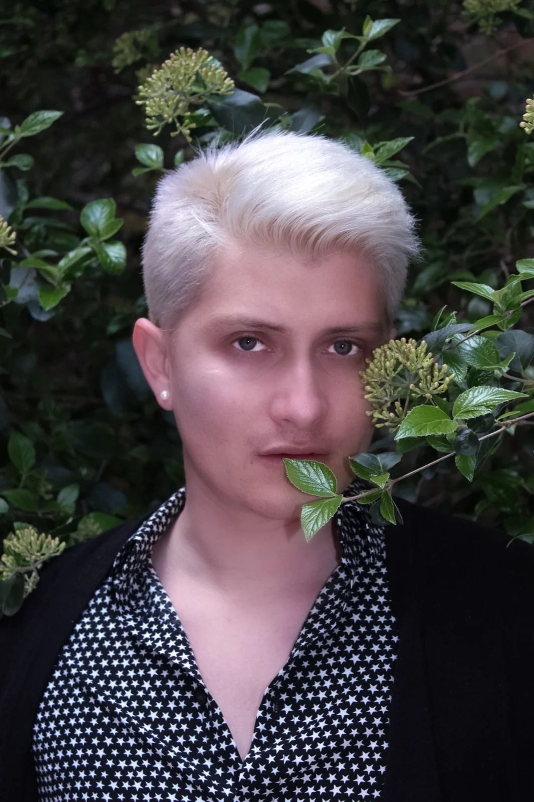 Person with short platinum hair in a black patterned shirt, holding a leaf near their mouth in a leafy outdoor setting