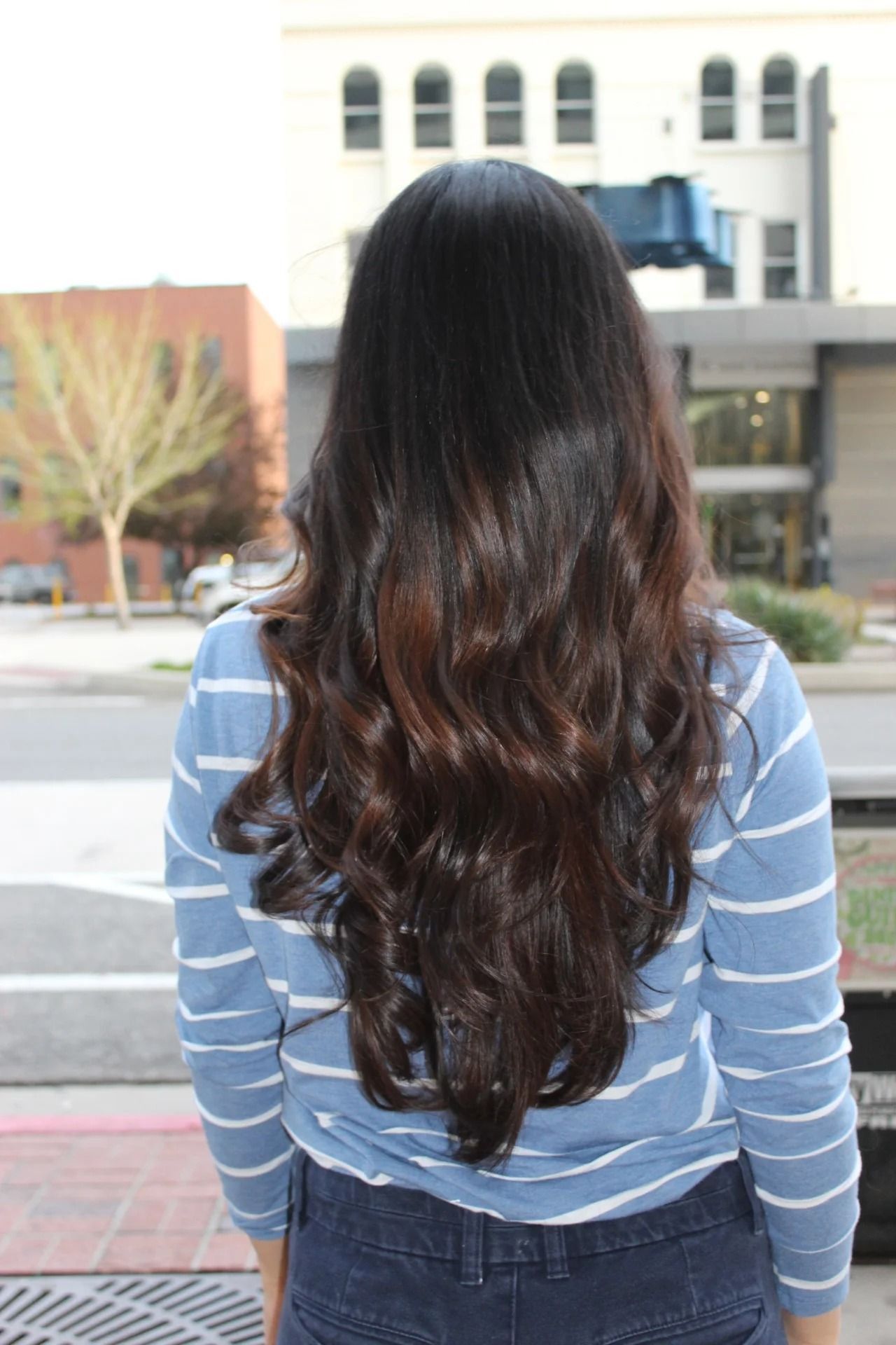 Person with long wavy dark hair, seen from behind, standing outdoors in a striped shirt near a building.
