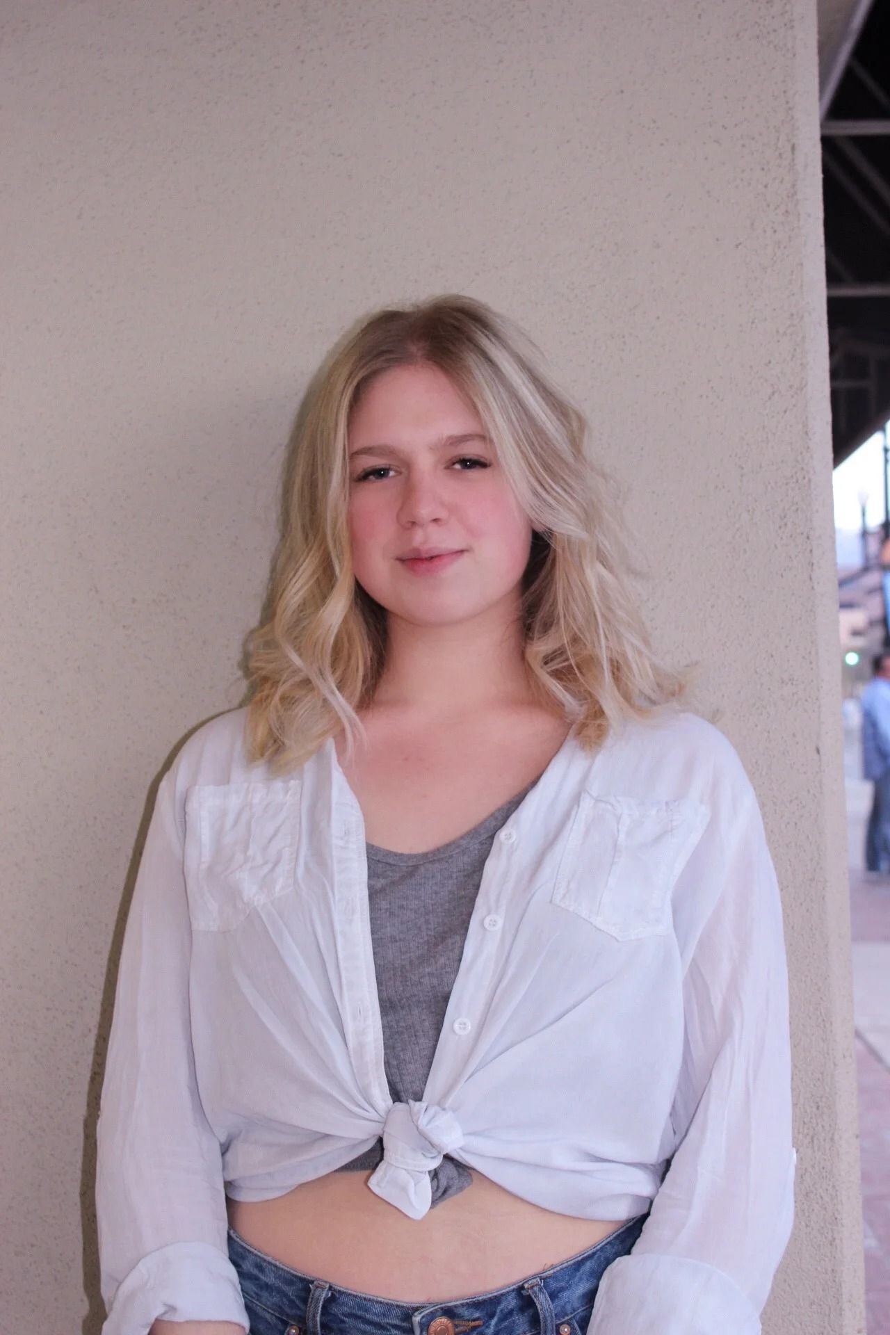 Woman in a white shirt and gray top standing against a wall, looking at the camera.