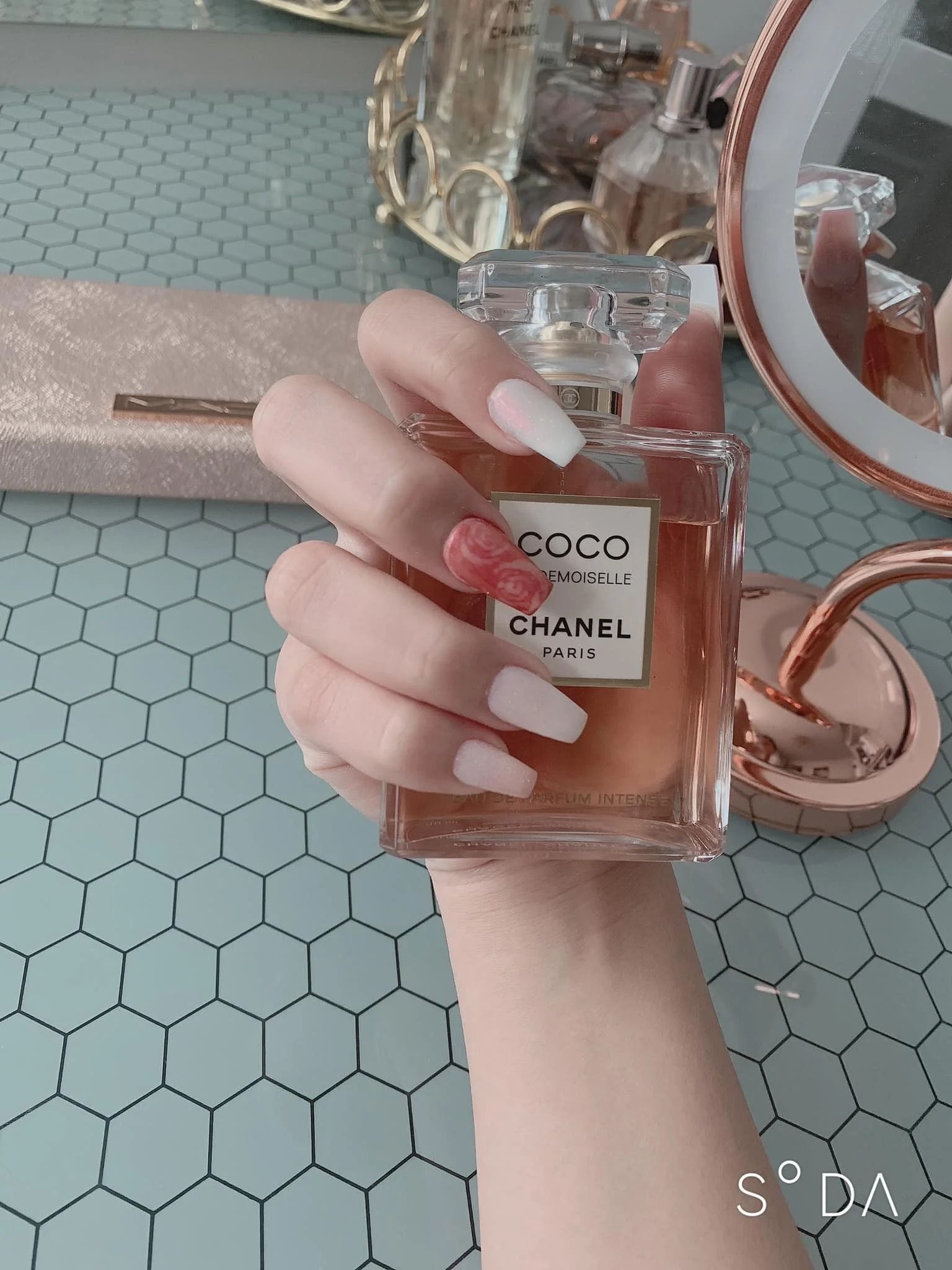 Hand holding a pink perfume bottle over a teal hexagon-tile surface and rose-gold mirror tray