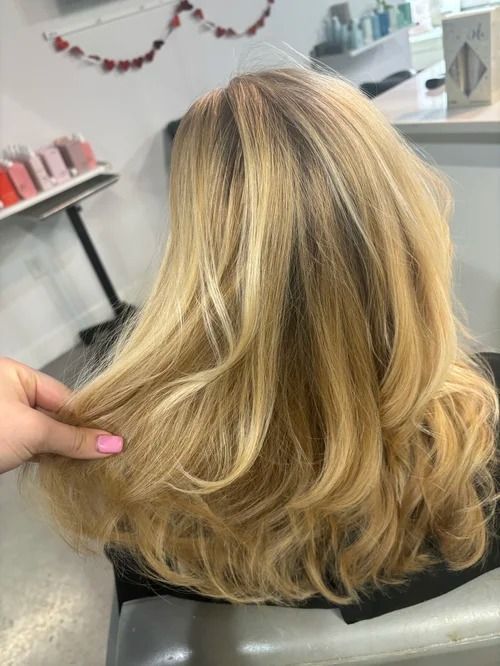Blonde shoulder-length wavy hair being held up in a salon chair with a hand showing pink nails