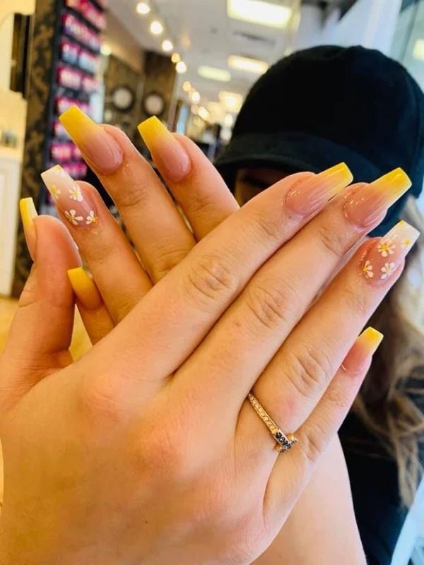 Hands with long yellow nails and glittery nail art held up in a salon, with a ring on one finger.