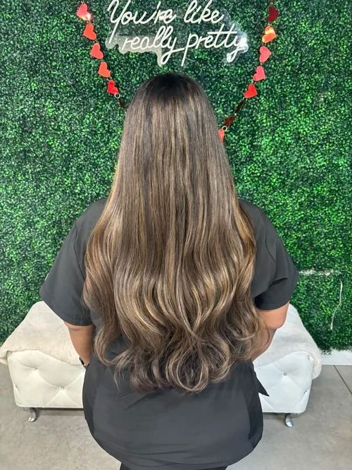 Woman with long wavy brown hair seated before a green wall with a heart and “You’re like really pretty” sign