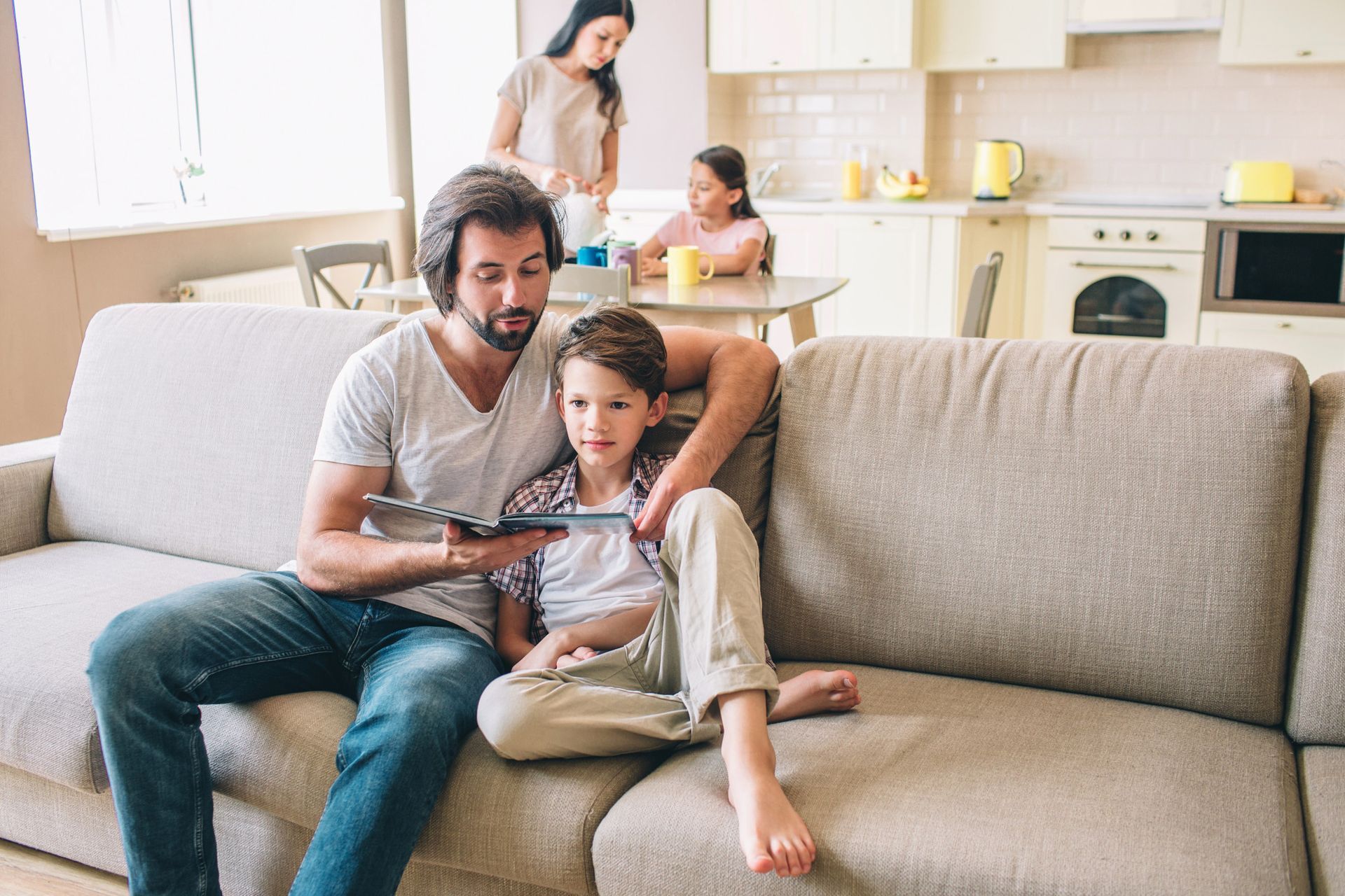 A parent and child sit on a couch reading a book together in a living room, with two other people in the kitchen behind.