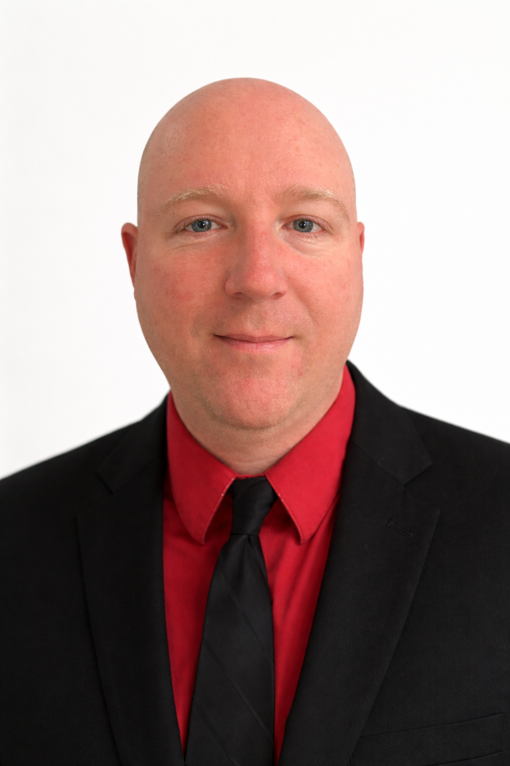 Studio headshot of a bald person in a black suit, red shirt, and black tie on a white background