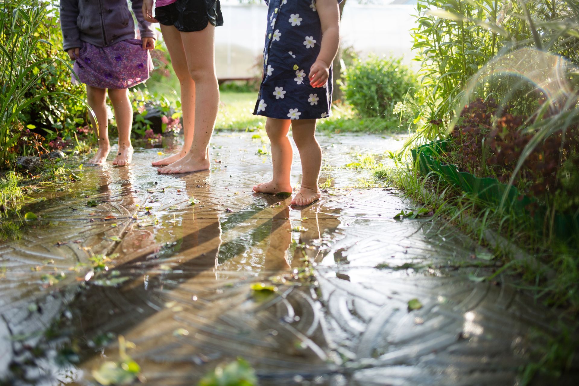 Three people stand barefoot on a wet stone path in a sunlit garden, surrounded by plants and lush greenery.