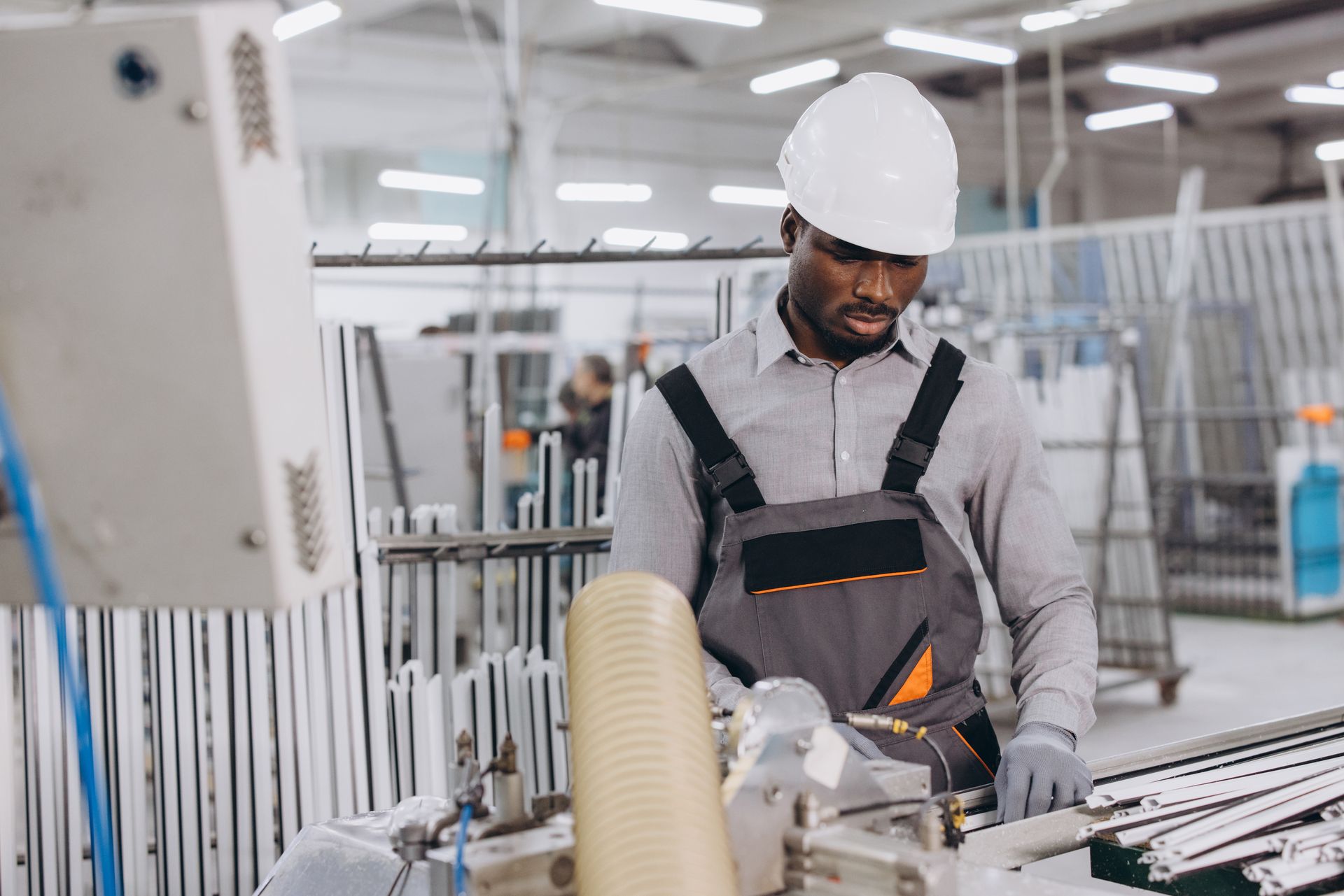 A worker in a white hard hat and grey overalls operates machinery in a bright industrial factory setting.