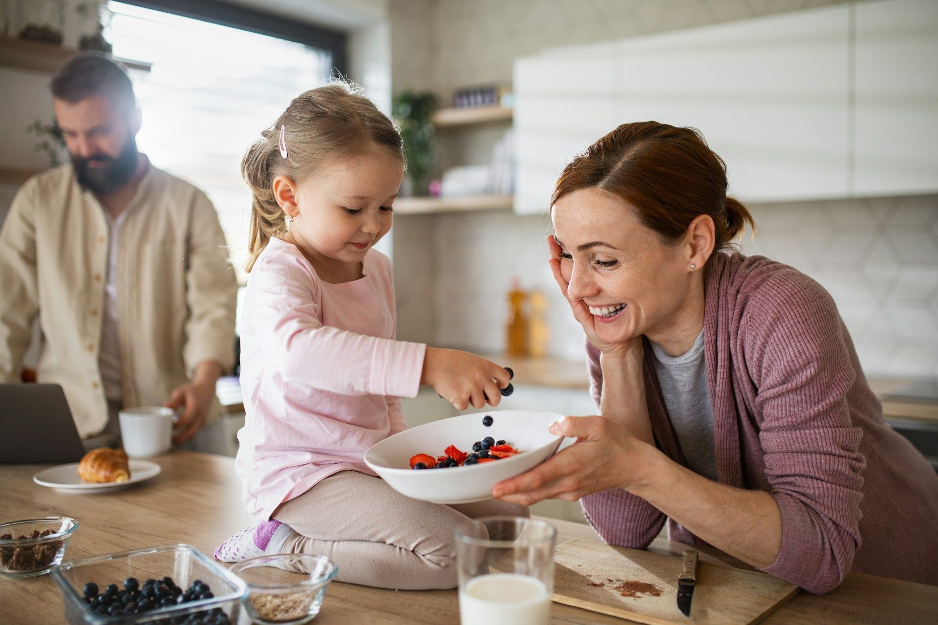 A child sitting on a kitchen counter places berries into a bowl held by an adult while another person works nearby.
