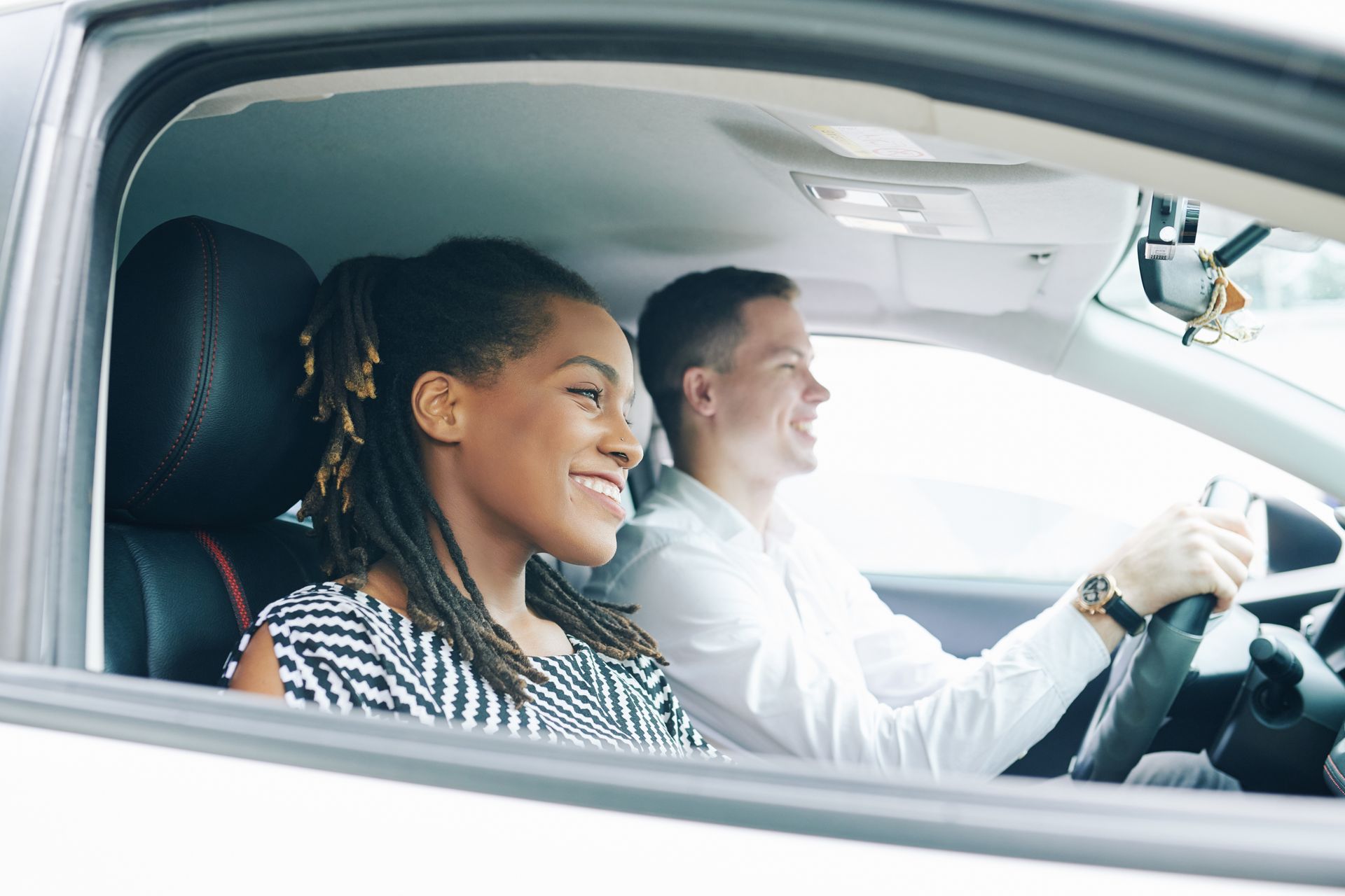 A person driving a car and a passenger in the front seat, both smiling while looking ahead.