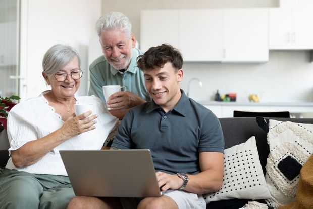 Three people on a couch looking at a laptop, one holding a cup and one reading a paper