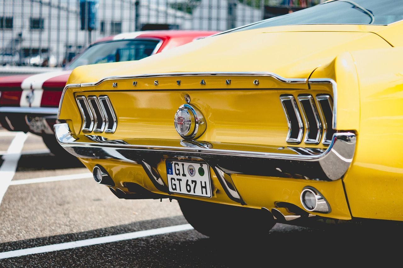 A bright yellow vintage Ford Mustang with a German license plate, parked in a lot behind a red car.