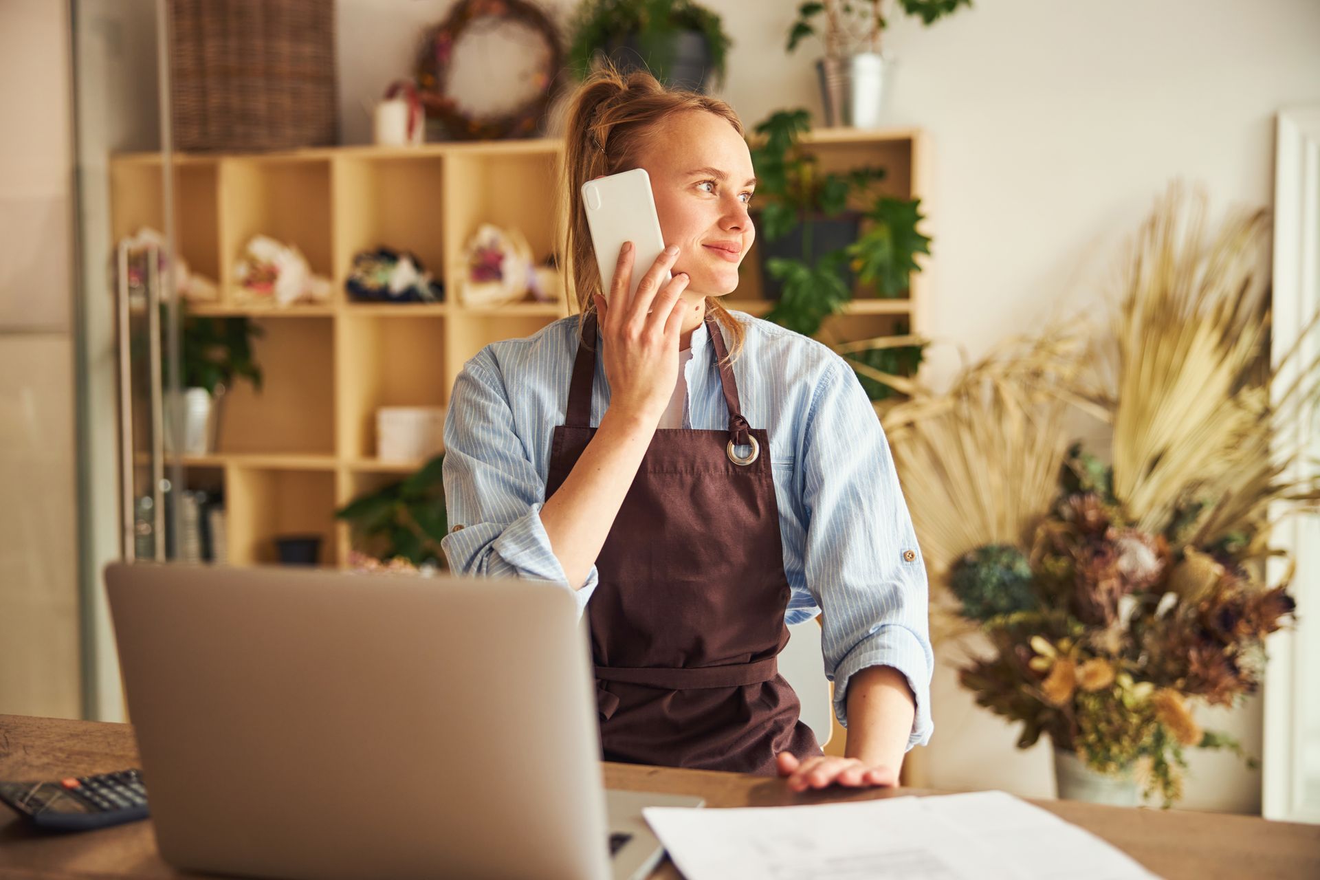 A person in an apron talks on a phone while working at a laptop in a shop filled with plants and decor.