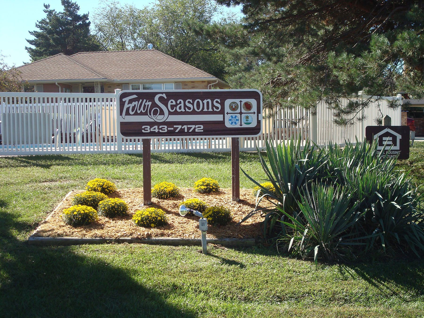 Sign for "Four Seasons" apartment complex with address and symbols, surrounded by landscaping.