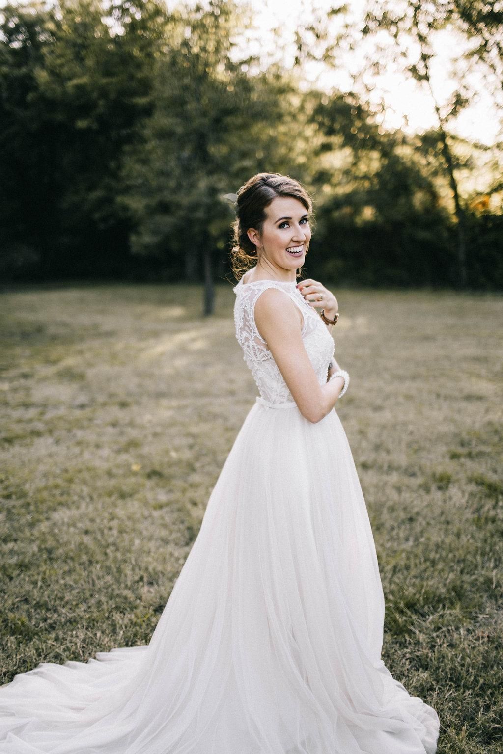 Bride in a white lace gown standing outdoors on a grassy lawn, smiling over her shoulder.