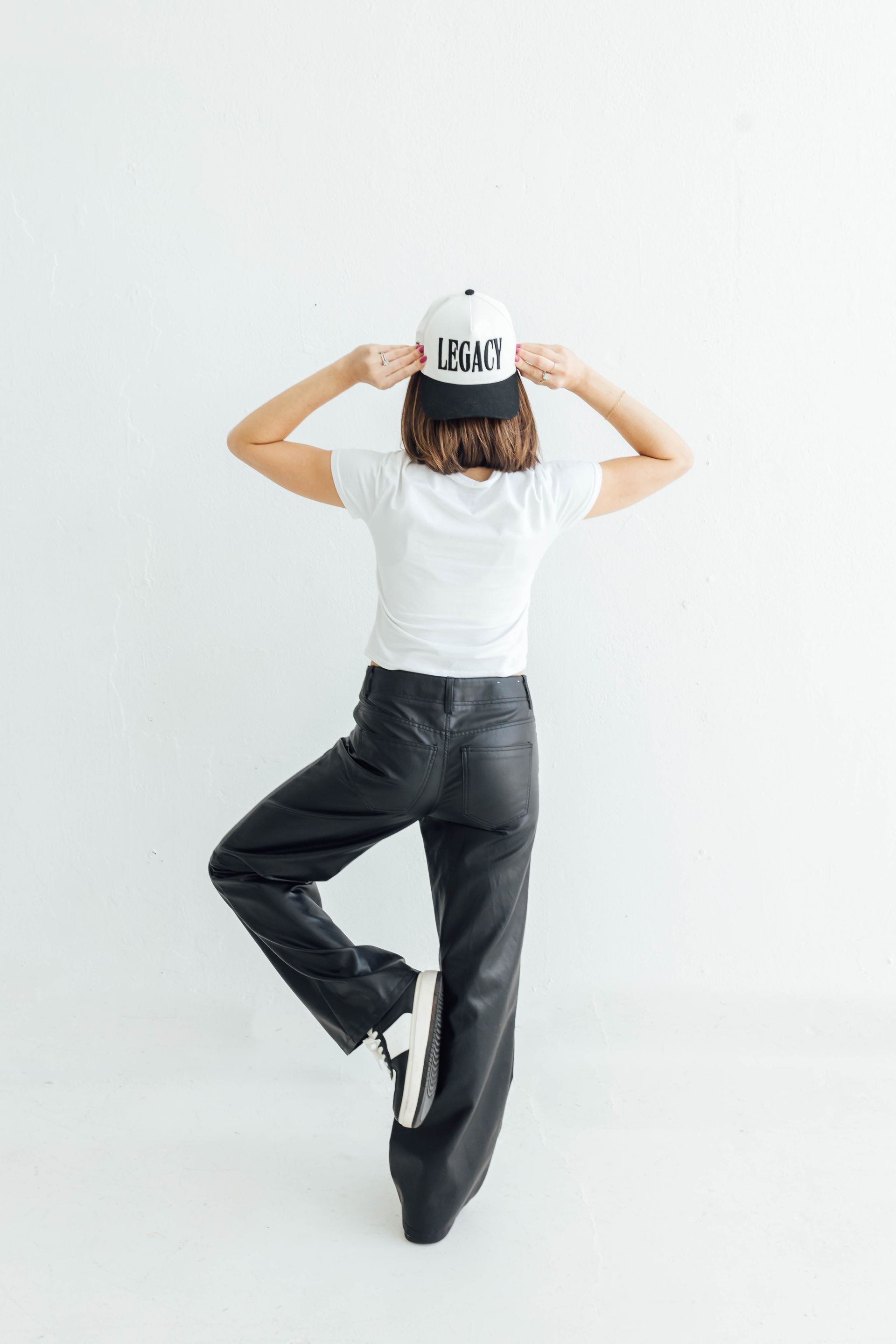 Person in white T-shirt and black pants posing against a white wall, adjusting a gray cap.