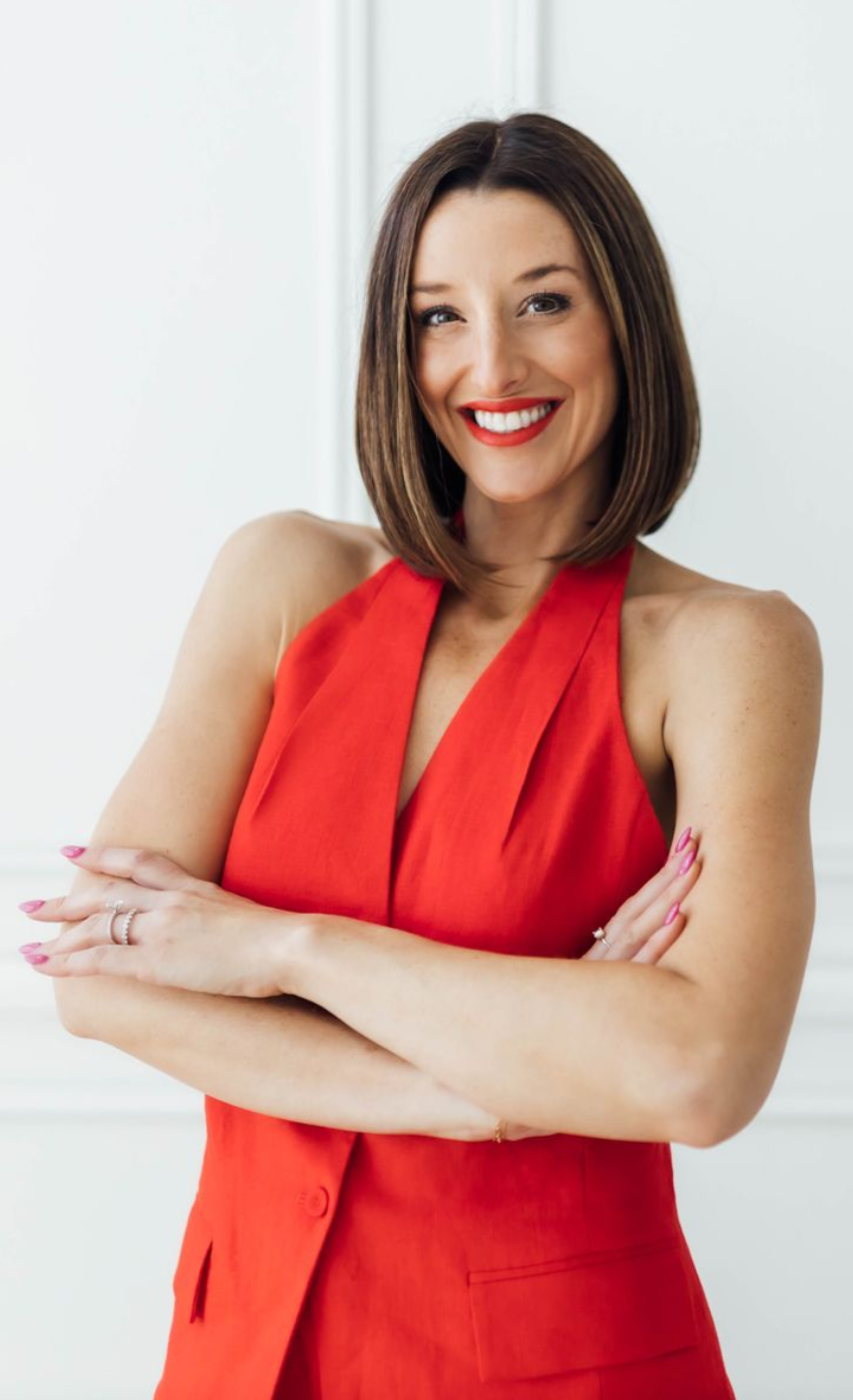 Smiling person in a red sleeveless blazer with arms crossed, standing against a light background