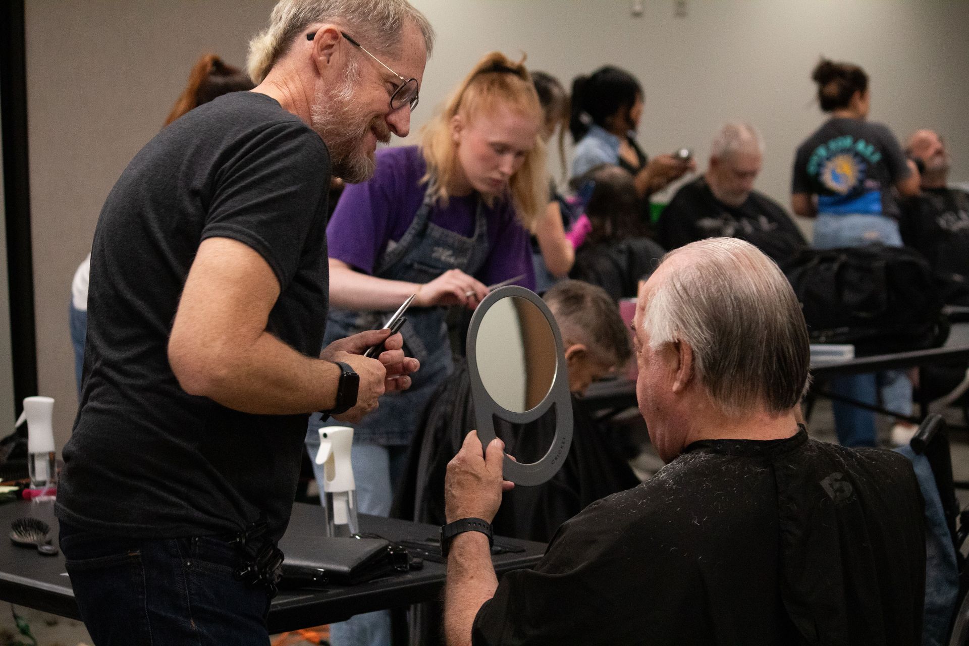 Two stylists working on a seated client in a busy salon, with mirrors and other people in the background