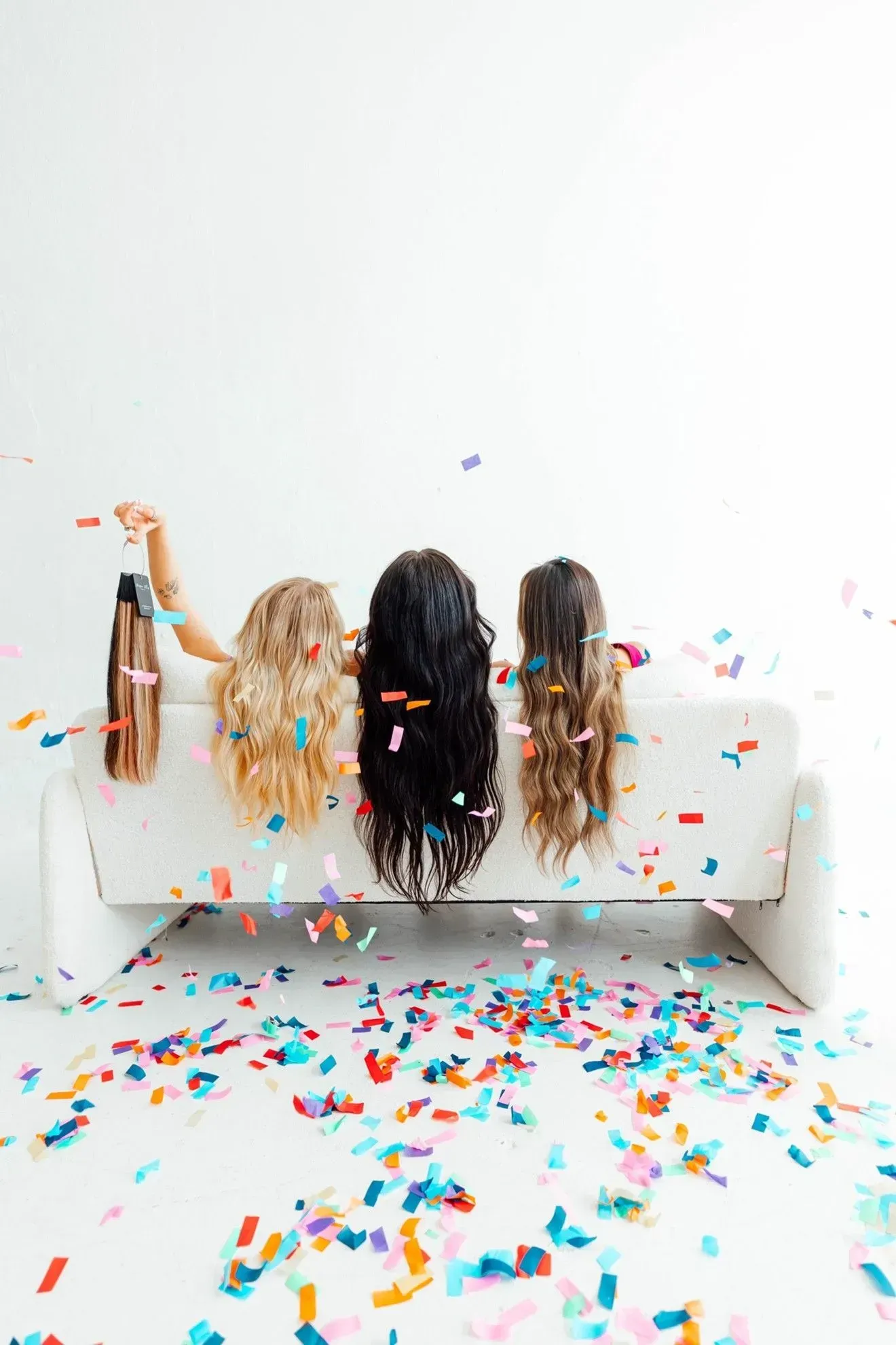 Three people on a white couch in a confetti-covered room, seen from behind, arms raised in celebration