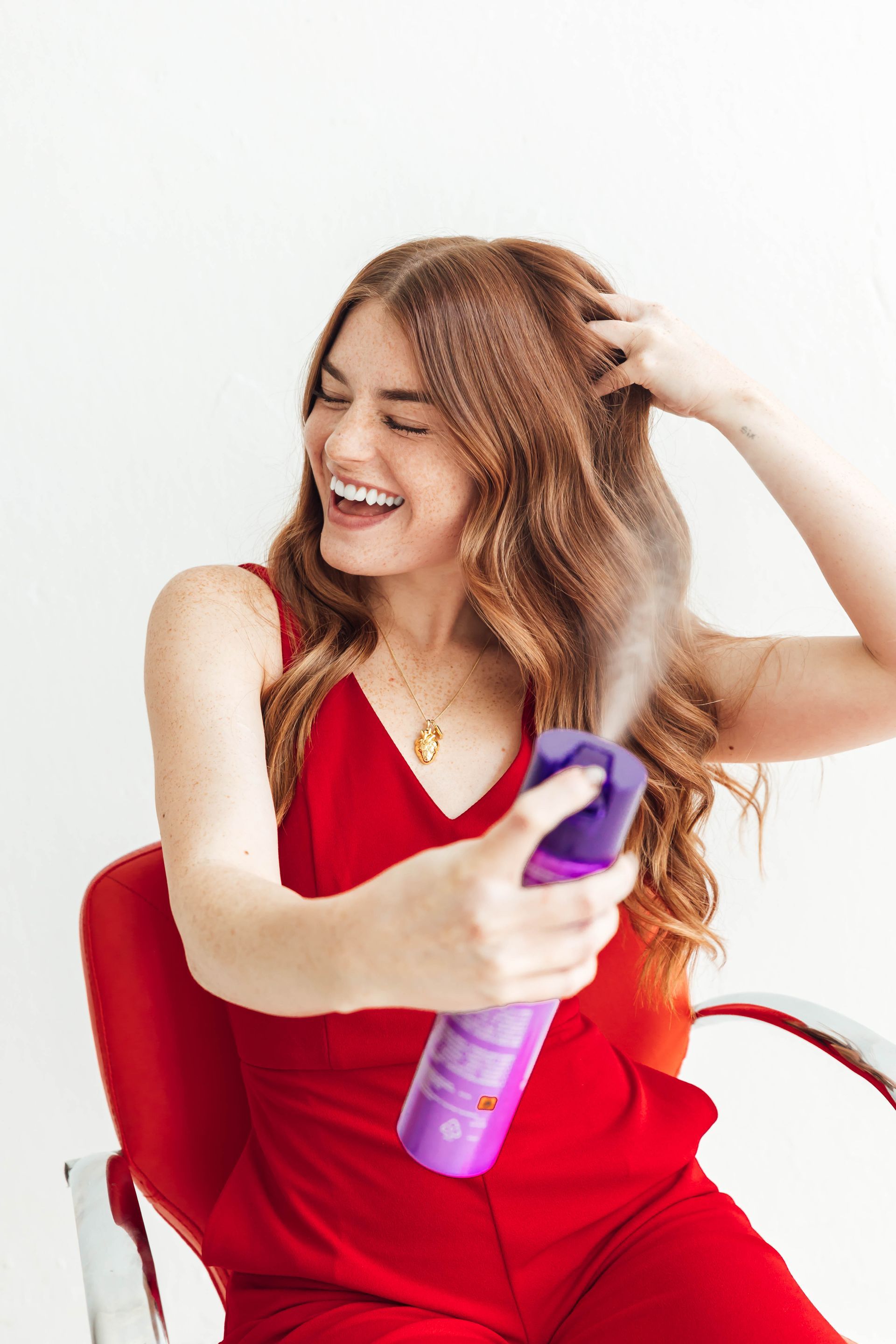 Woman in a red dress laughing while spraying purple hairspray against a white background