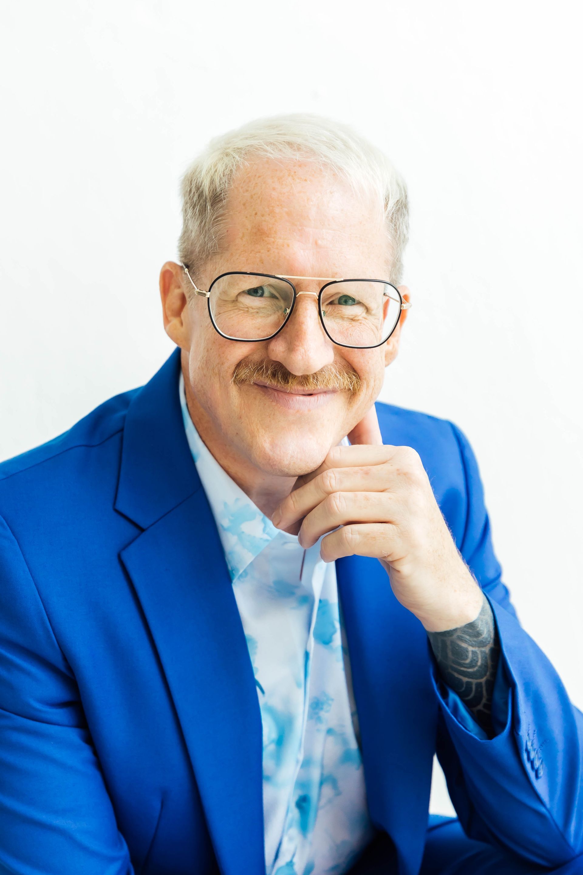Smiling man in a bright blue blazer, resting his chin on his hand against a white background