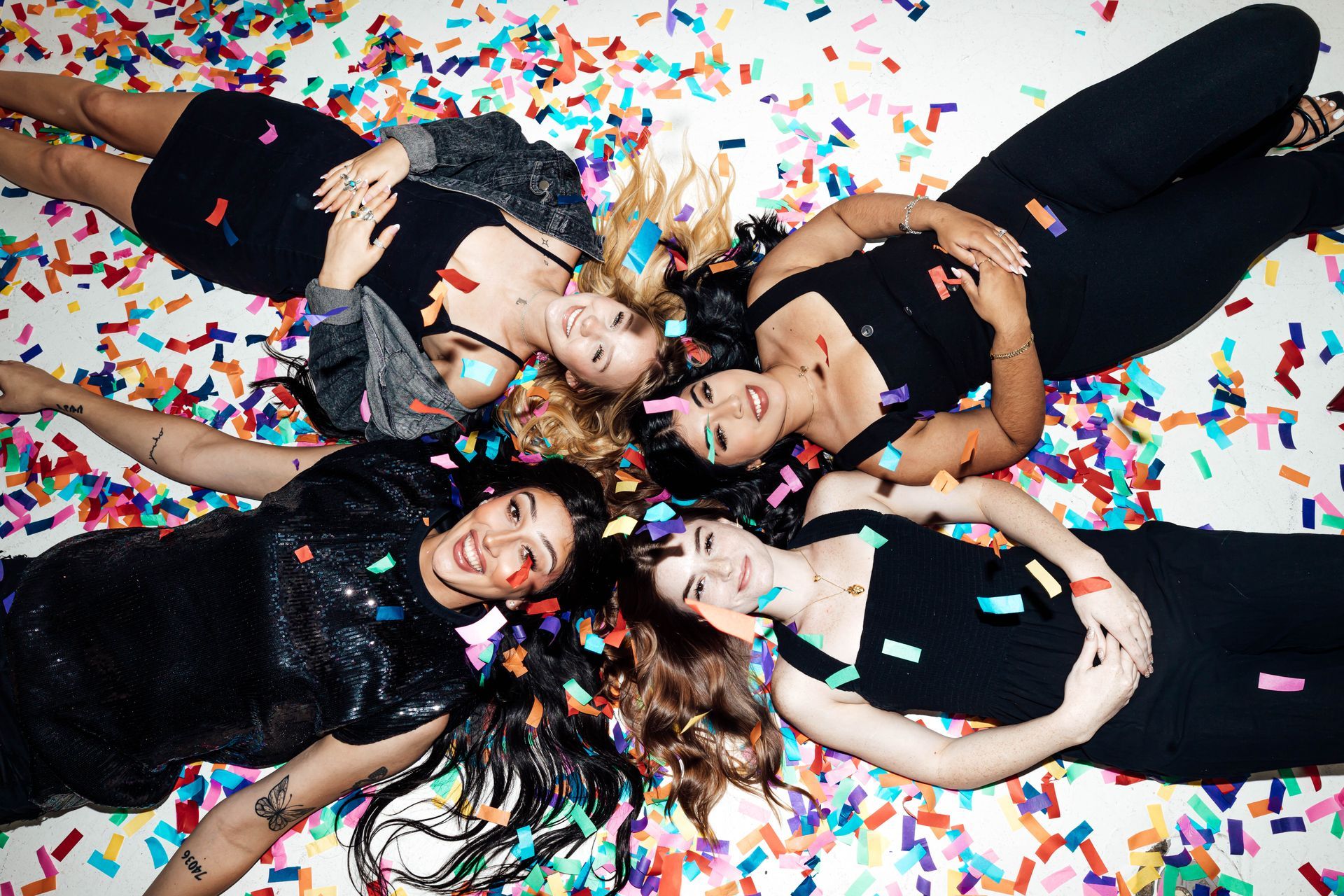 Five friends lying in a circle on confetti-covered floor, smiling at the camera