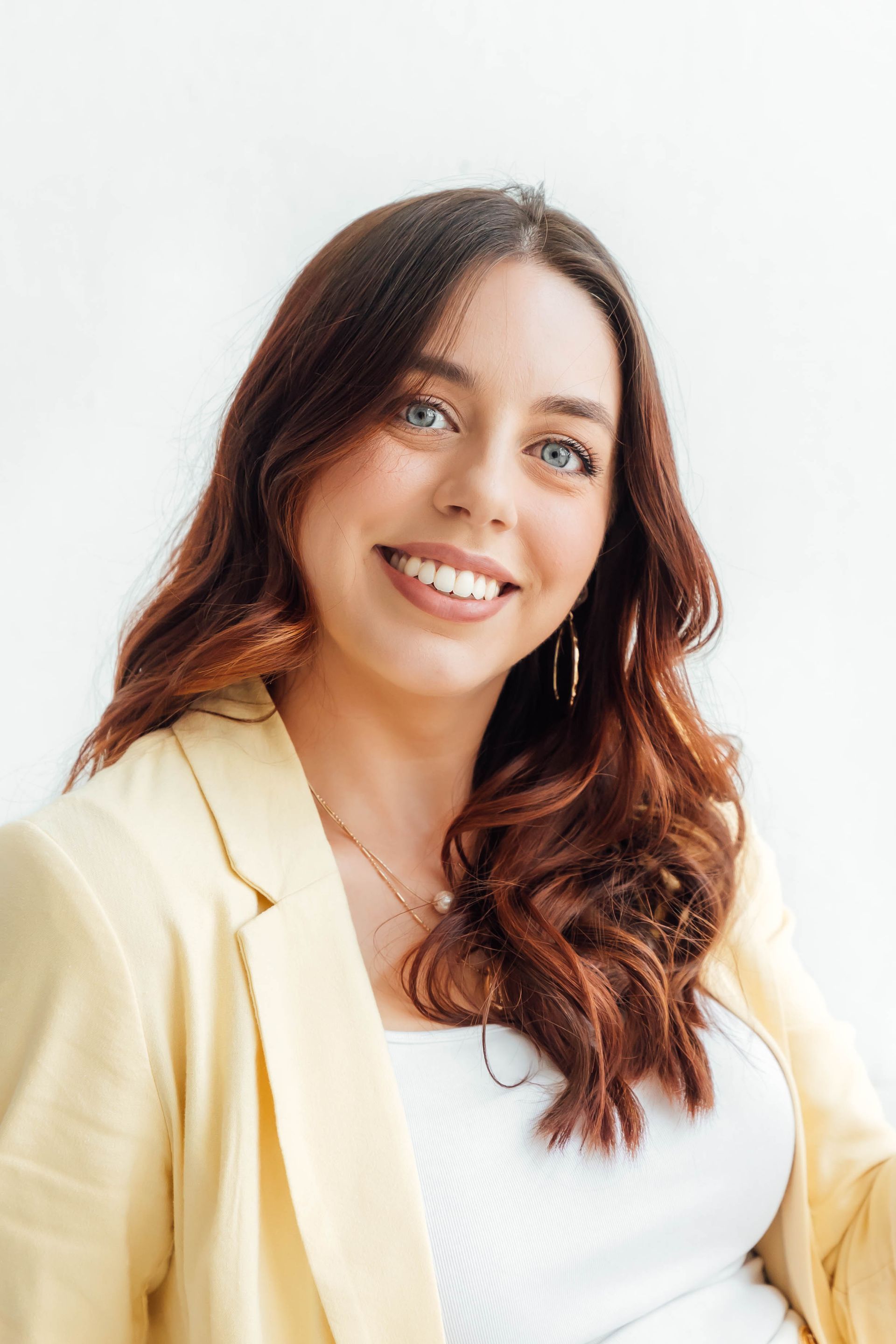 Smiling woman with curly brown hair wearing a light yellow blazer and white top against a white background