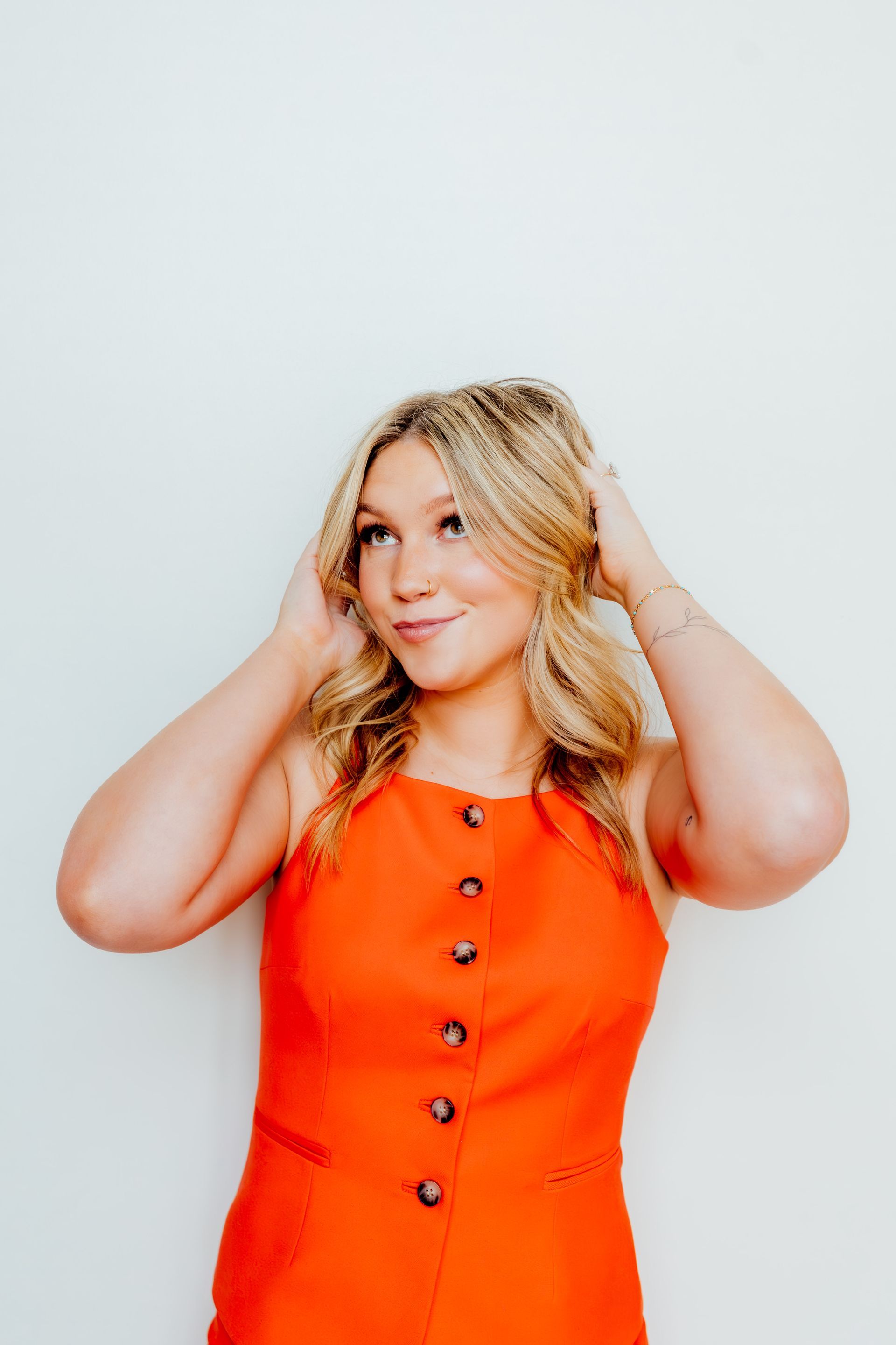 Person in an orange button-front dress, smiling and touching their hair against a white background