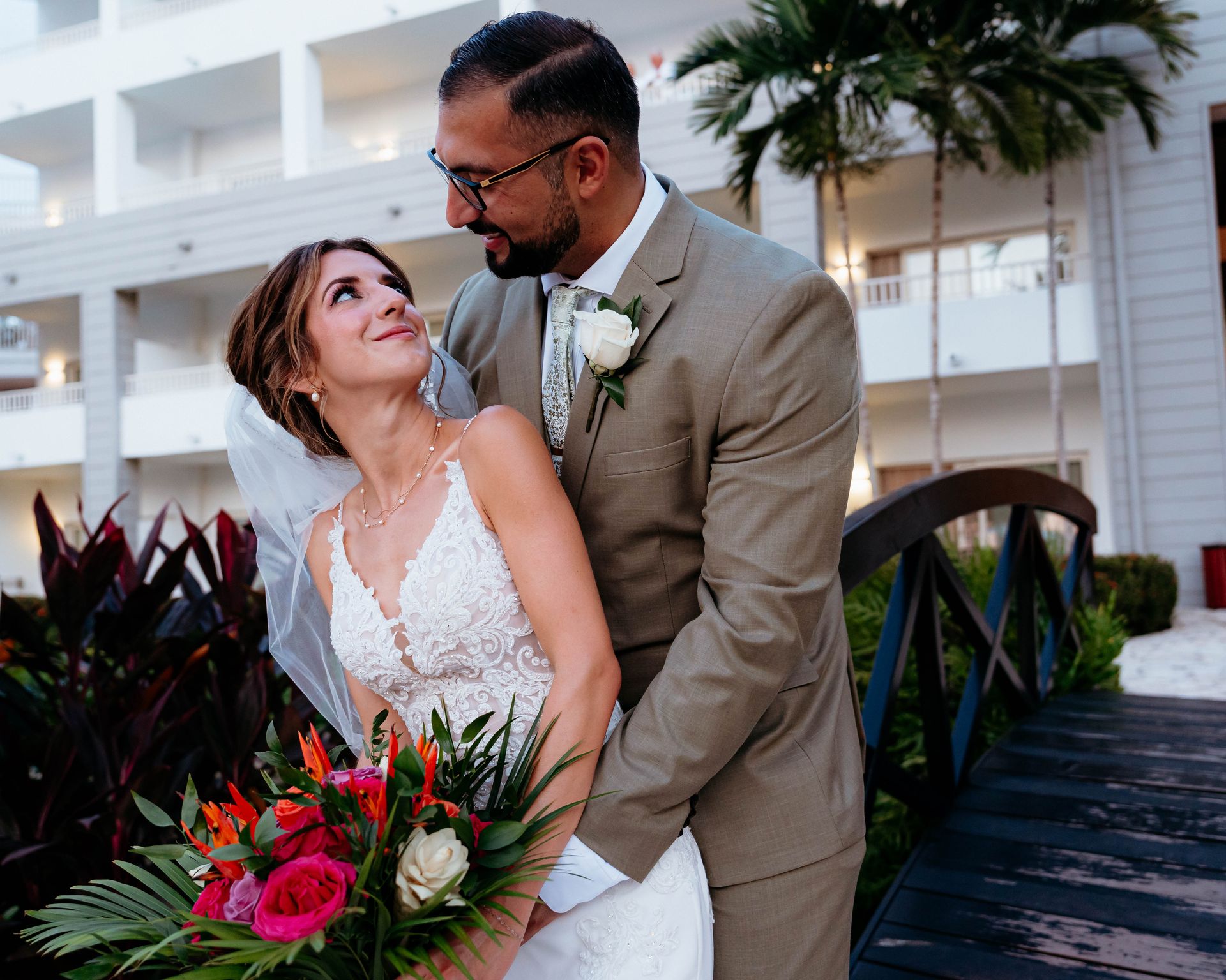 Bride and groom smiling on a bridge at a wedding, with tropical flowers and palm trees nearby