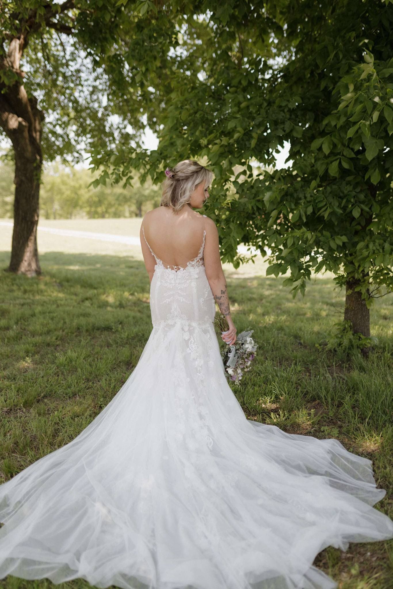 Bride in a white wedding gown standing outdoors beneath trees, with a long flowing train.
