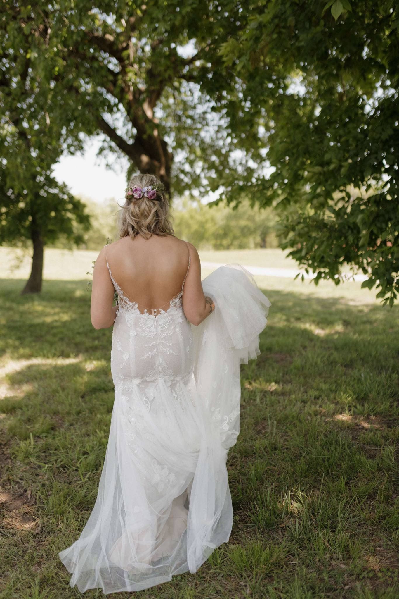 Bride in a white lace gown walking beneath trees in a grassy field