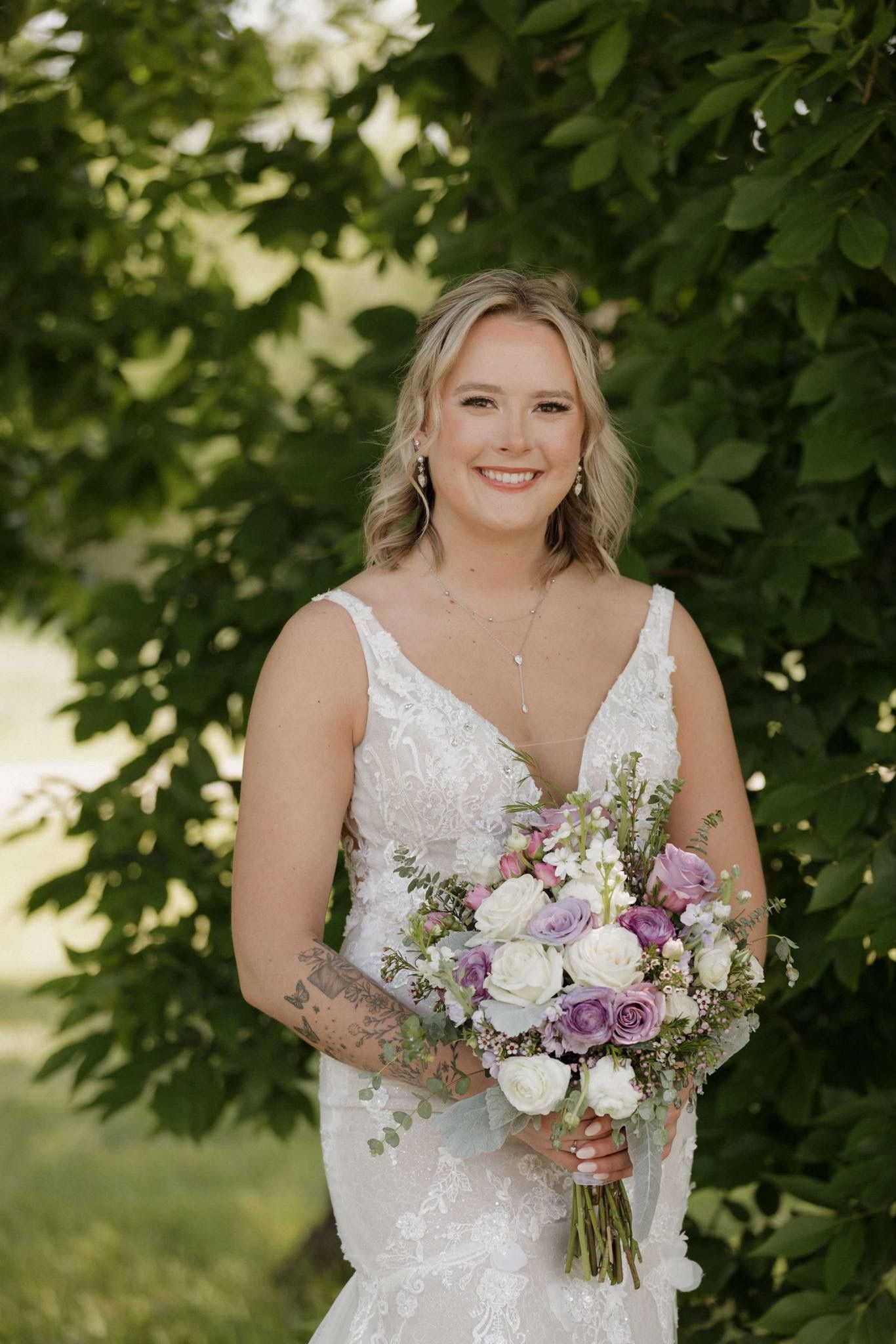 Bride in a white lace dress holding a purple and white bouquet outdoors under green trees
