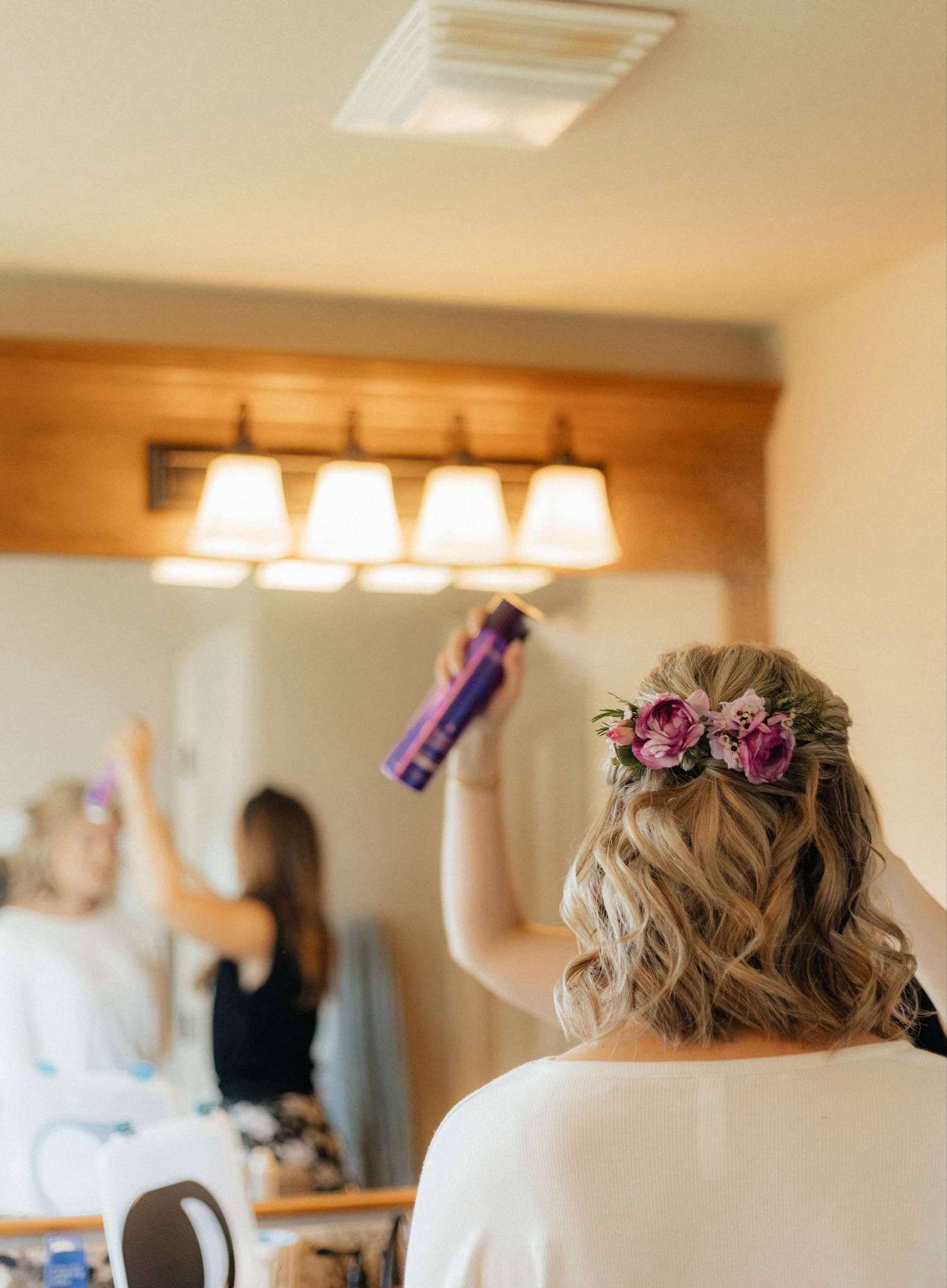 Woman with floral hair accessory holding hairspray in a bathroom mirror, preparing her hairstyle.