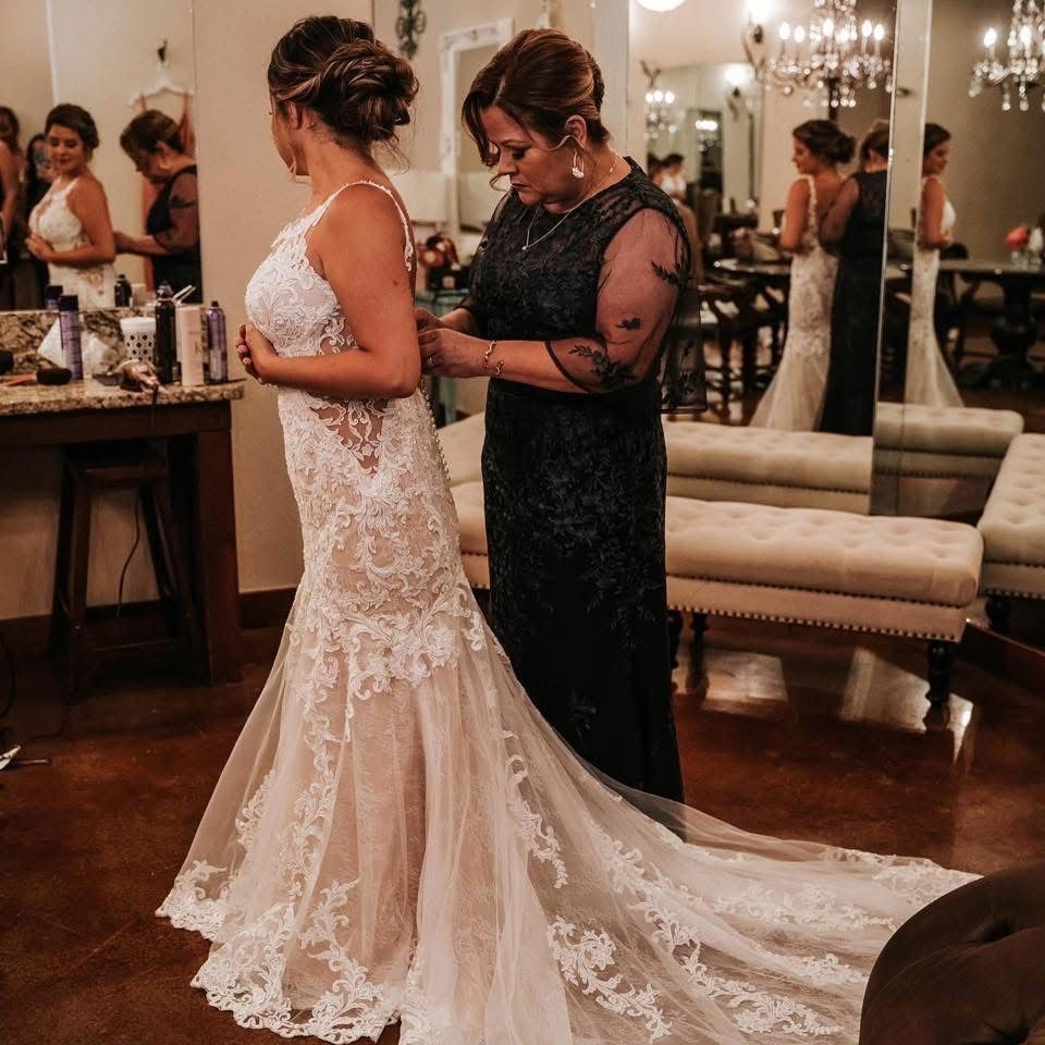 Bride being fastened into a lace wedding gown by a helper in a formal room