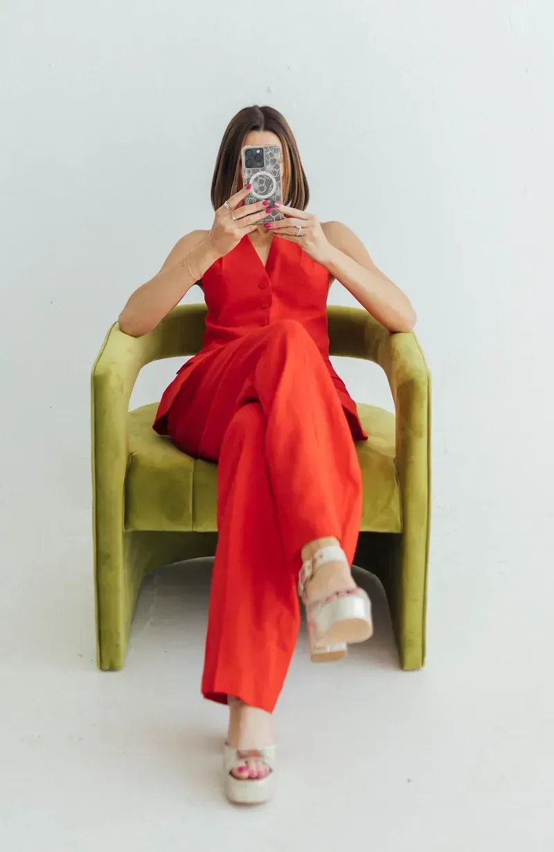 Person in a red outfit sitting in a green chair, taking a mirror selfie against a white backdrop.