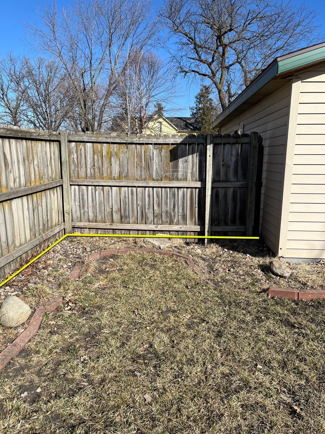 Backyard corner with wood fence, black gate, yellow measuring tape, and house siding.