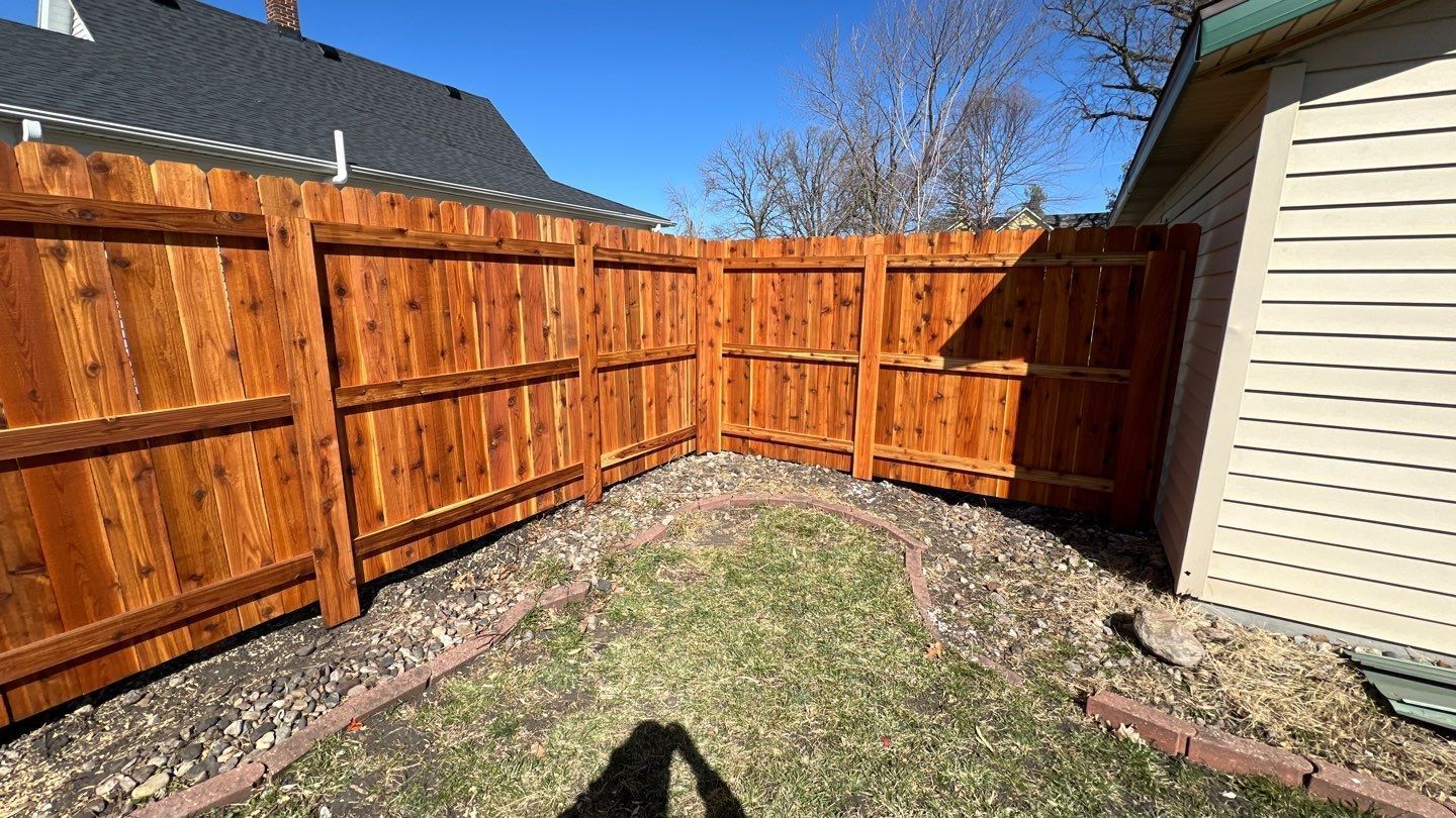 Wooden privacy fence encloses a gravel backyard beside a beige house in bright sunlight.