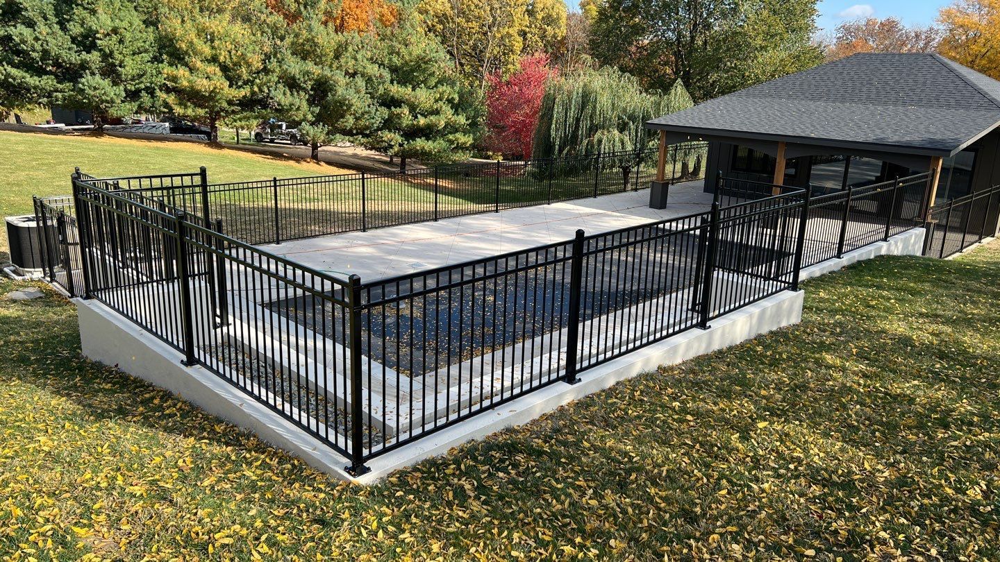 Black metal fenced patio with covered hot tub beside a pavilion, surrounded by autumn trees and grass.