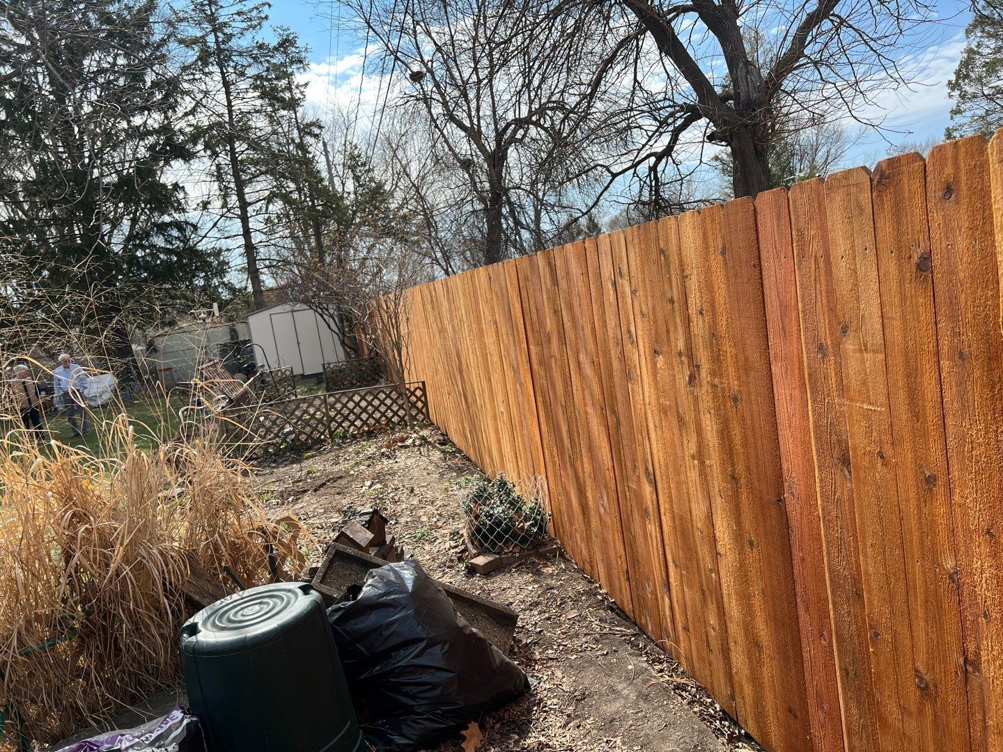 Backyard with a tall wooden fence, trash bins, and dry brush under bare trees