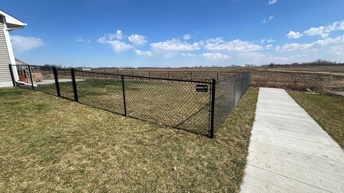 Black chain-link fence along a grassy field beside a concrete path under a blue sky.