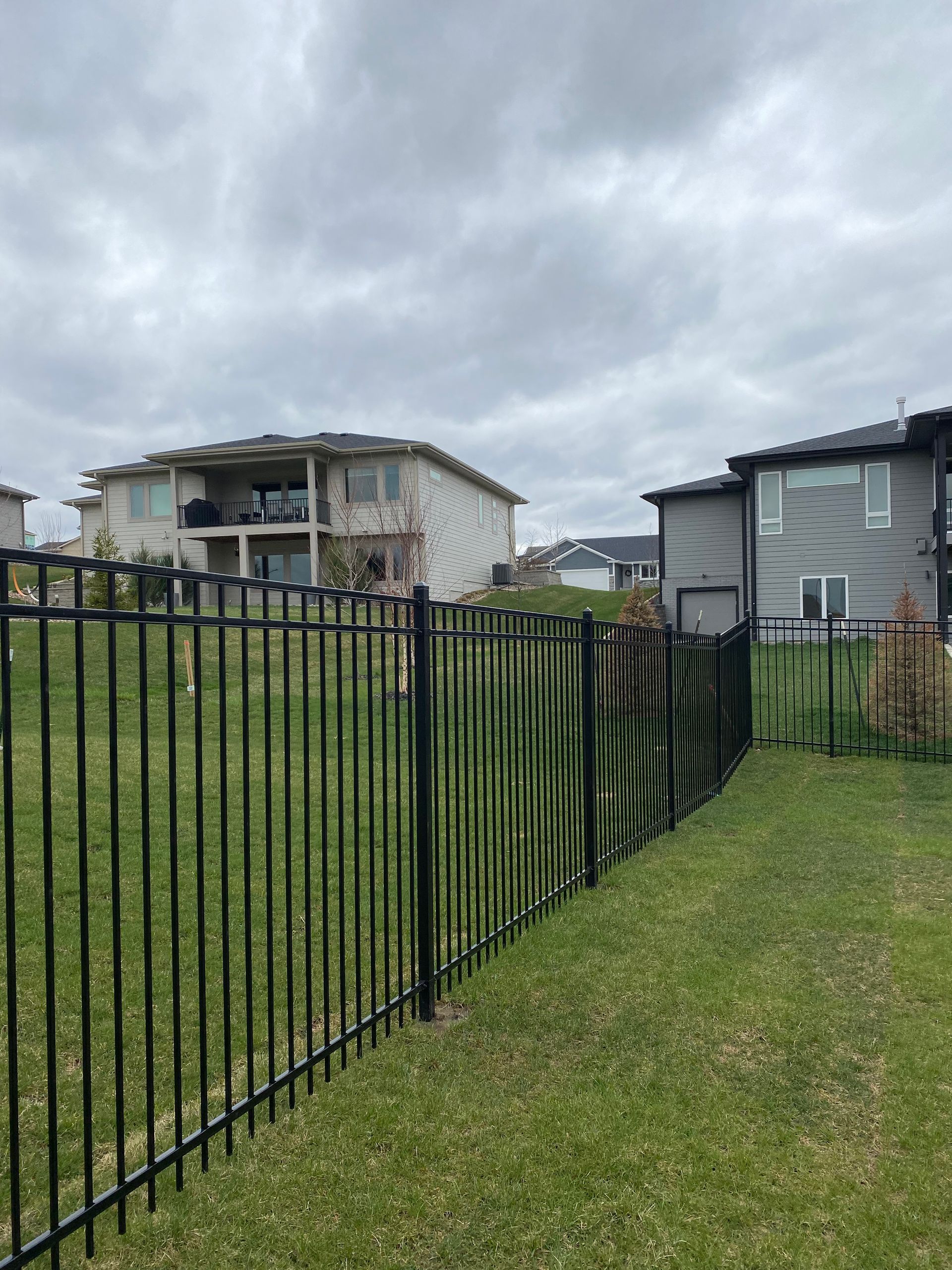 Black metal fence along a grassy yard beside damaged houses under a cloudy sky.