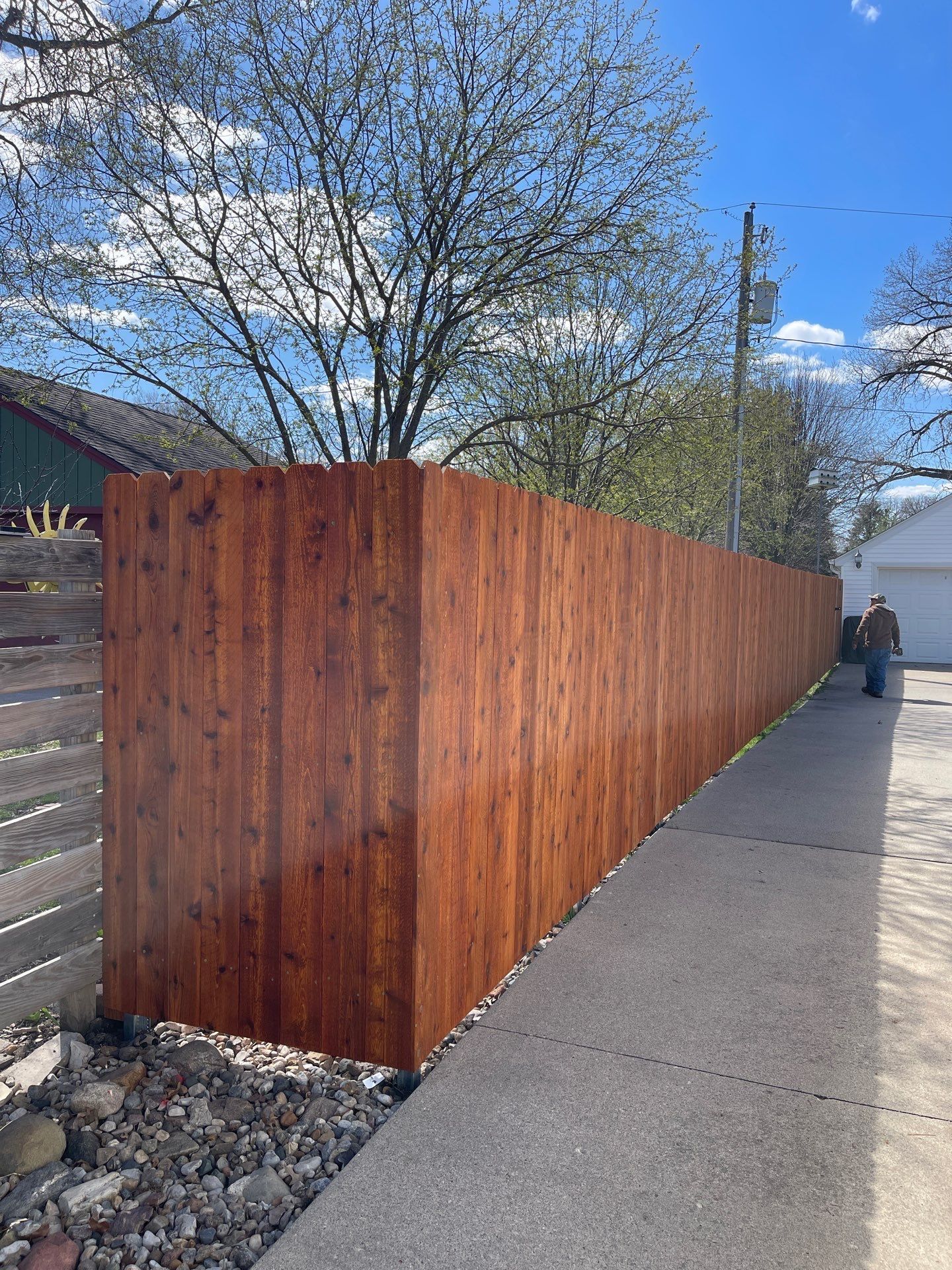 Long wooden fence lining a sidewalk, with a person walking in the distance under a blue sky.