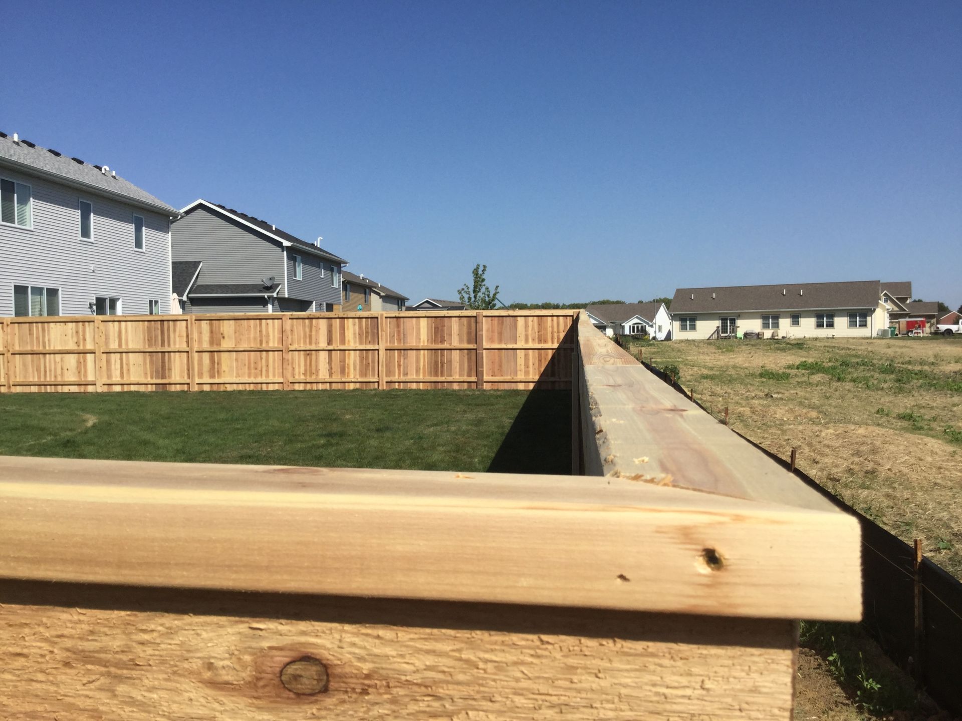 Residential backyard with a wooden fence, grassy trench, and concrete retaining wall under a blue sky