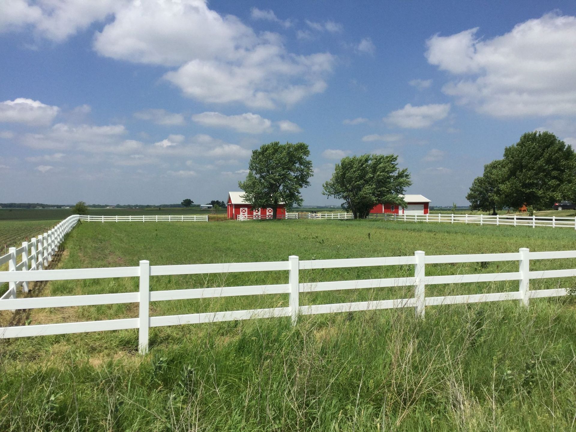 White fence around green field with red barns and trees under a blue sky