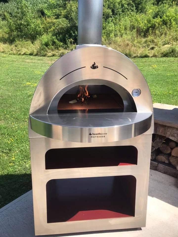 Stainless steel outdoor pizza oven with fire burning inside. Green grass and sky in the background.