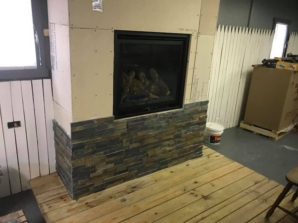 Fireplace under construction, partially tiled with stacked stone. Drywall, wood paneling, and flooring are visible.