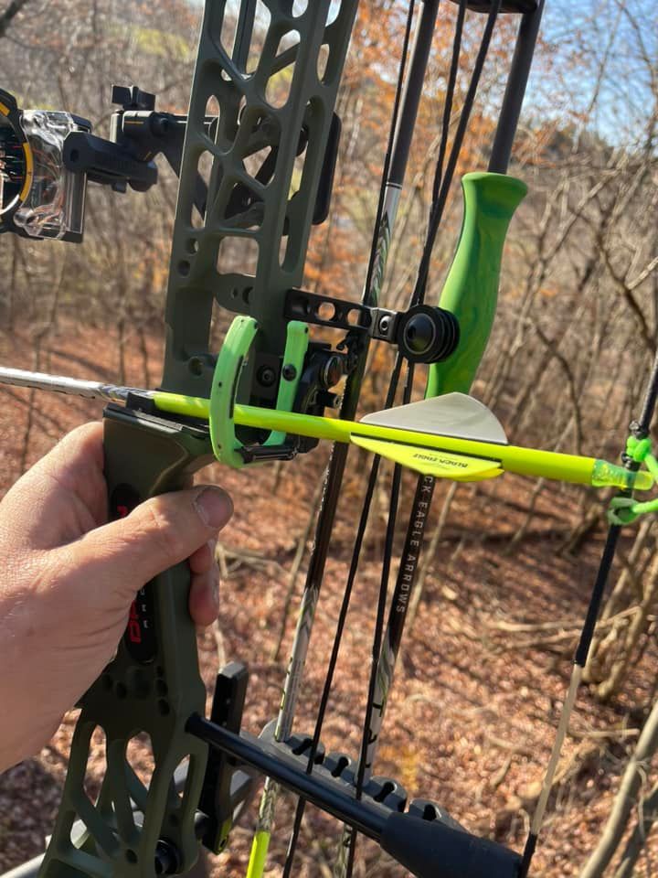 A hand holding a green compound bow with a fluorescent yellow arrow. Outdoors in a wooded area.