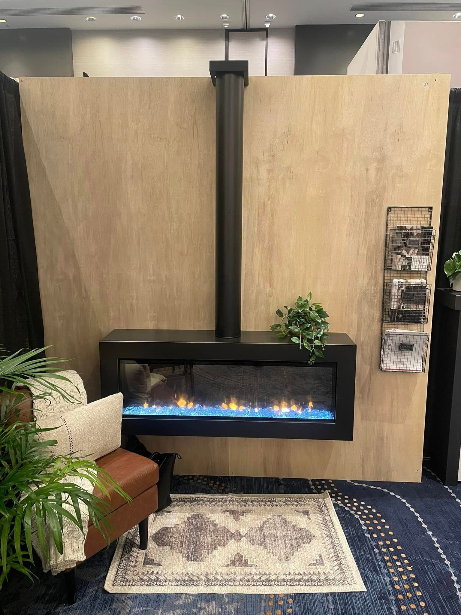 Modern fireplace with blue flames, black frame and chimney, on beige wall. Decorative rug and plants in front.