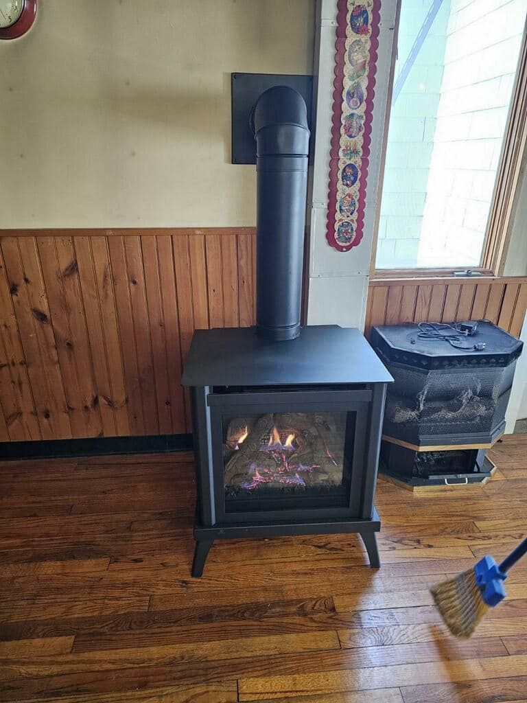 Black fireplace with lit flames, black chimney pipe, on wood floor, near a window.