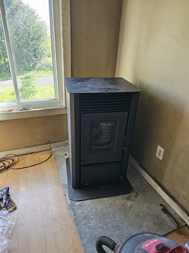 Pellet stove in corner of a room, next to a window. Black stove on a dark mat, tan walls.