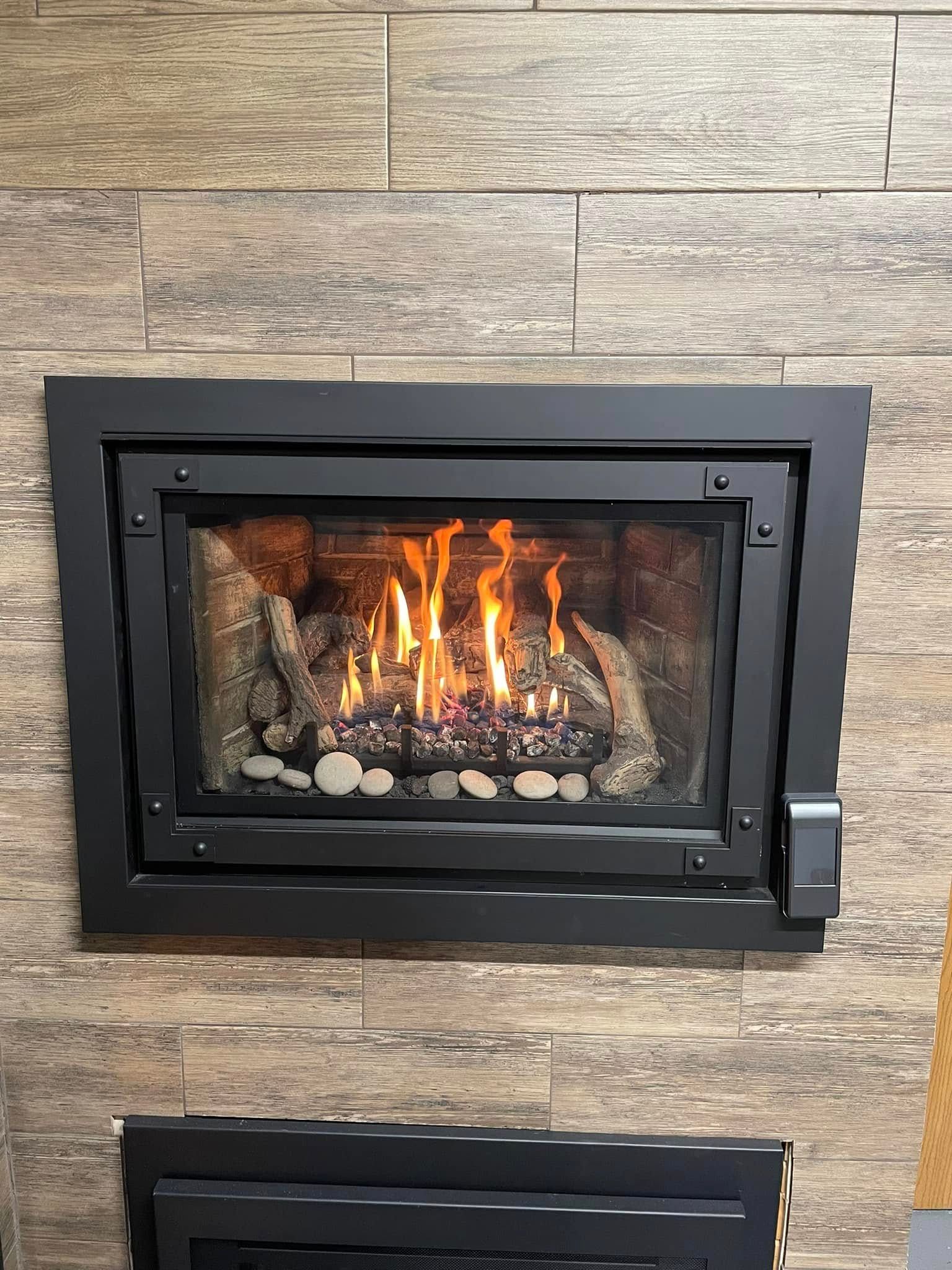 A gas fireplace with a black frame and burning flames in a stone-lined enclosure, mounted on a wood-look wall.